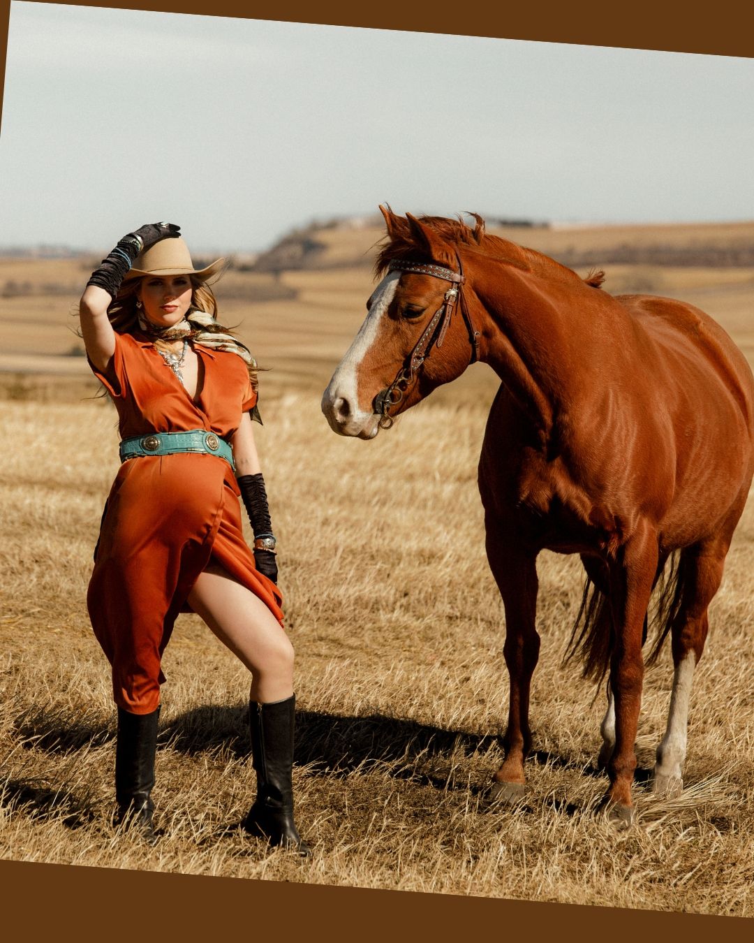 A brown horse stands in a dry field next to someone wearing a western-style orange outfit and cowboy hat.