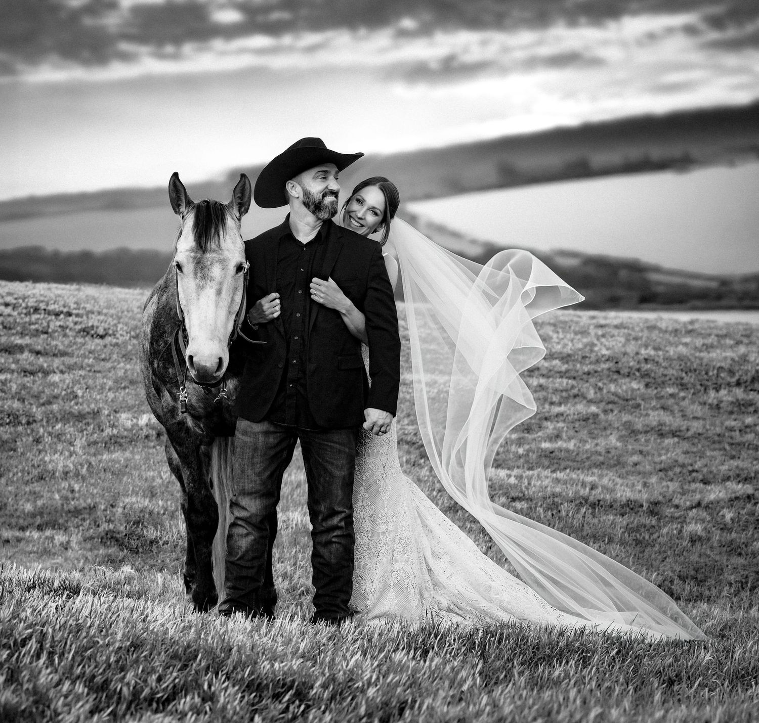 Black and white wedding photo of a couple in western attire standing with a horse in an open field.