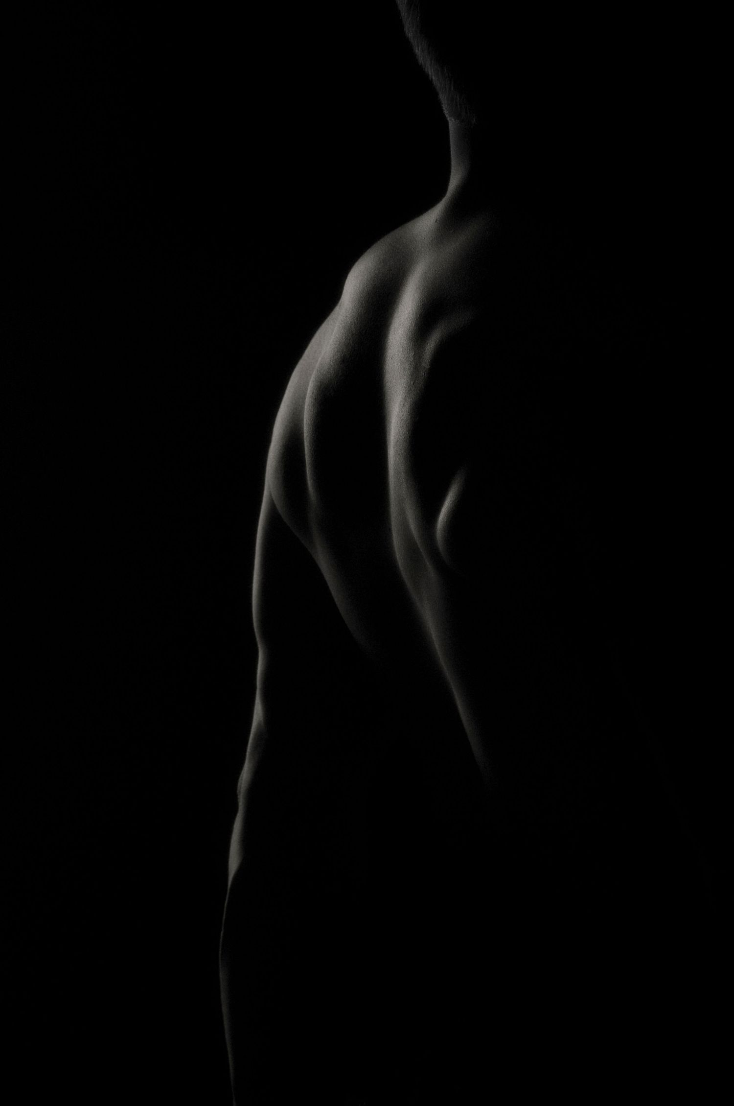 Dramatic low-key artistic photo of a muscular male back, highlighted by chiaroscuro lighting against a dark background.