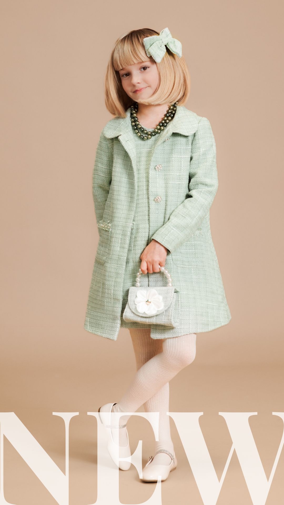 Girl in mint green tweed coat dress with pearl necklace, bow hair clip, and silver handbag, posing for new collection.