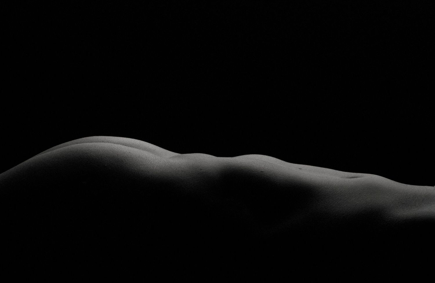 Black and white fine art nude photograph showcasing the curves of a human body against a dark background.