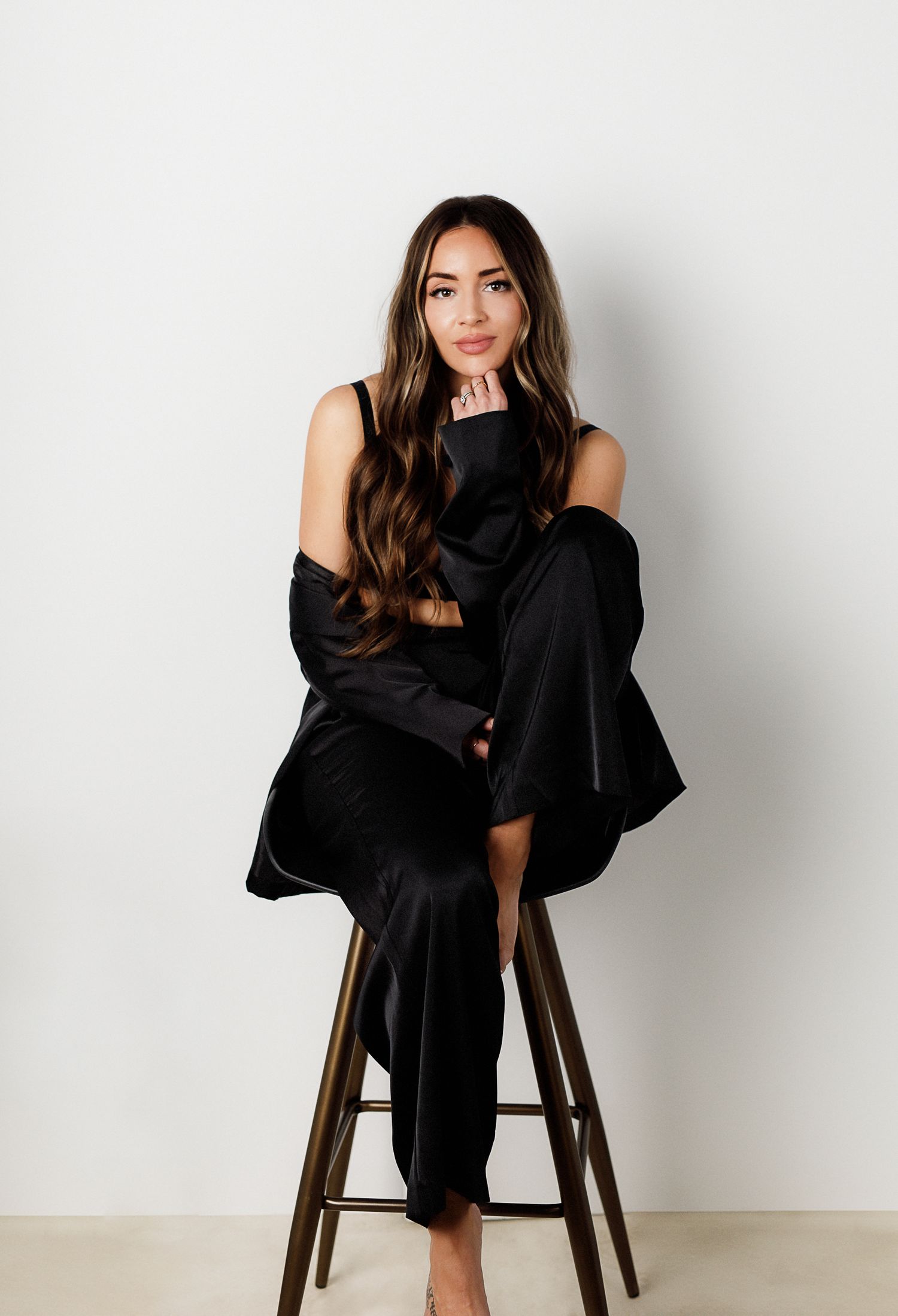 Minimalist portraits in all black attire seated on wooden stool against white background.