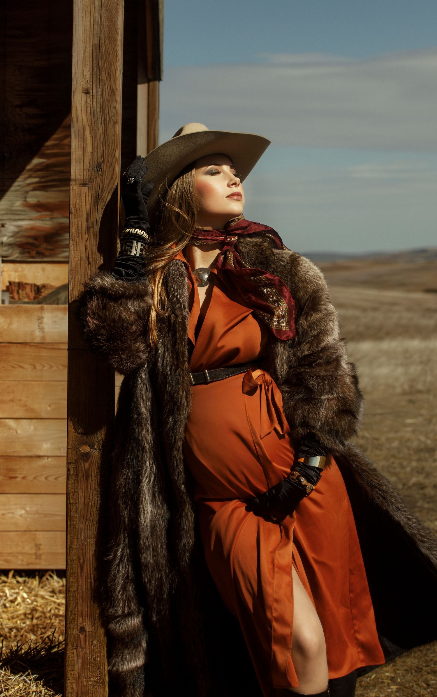A western-styled model in an orange dress and fur coat poses against a rustic wooden building in a desert landscape.