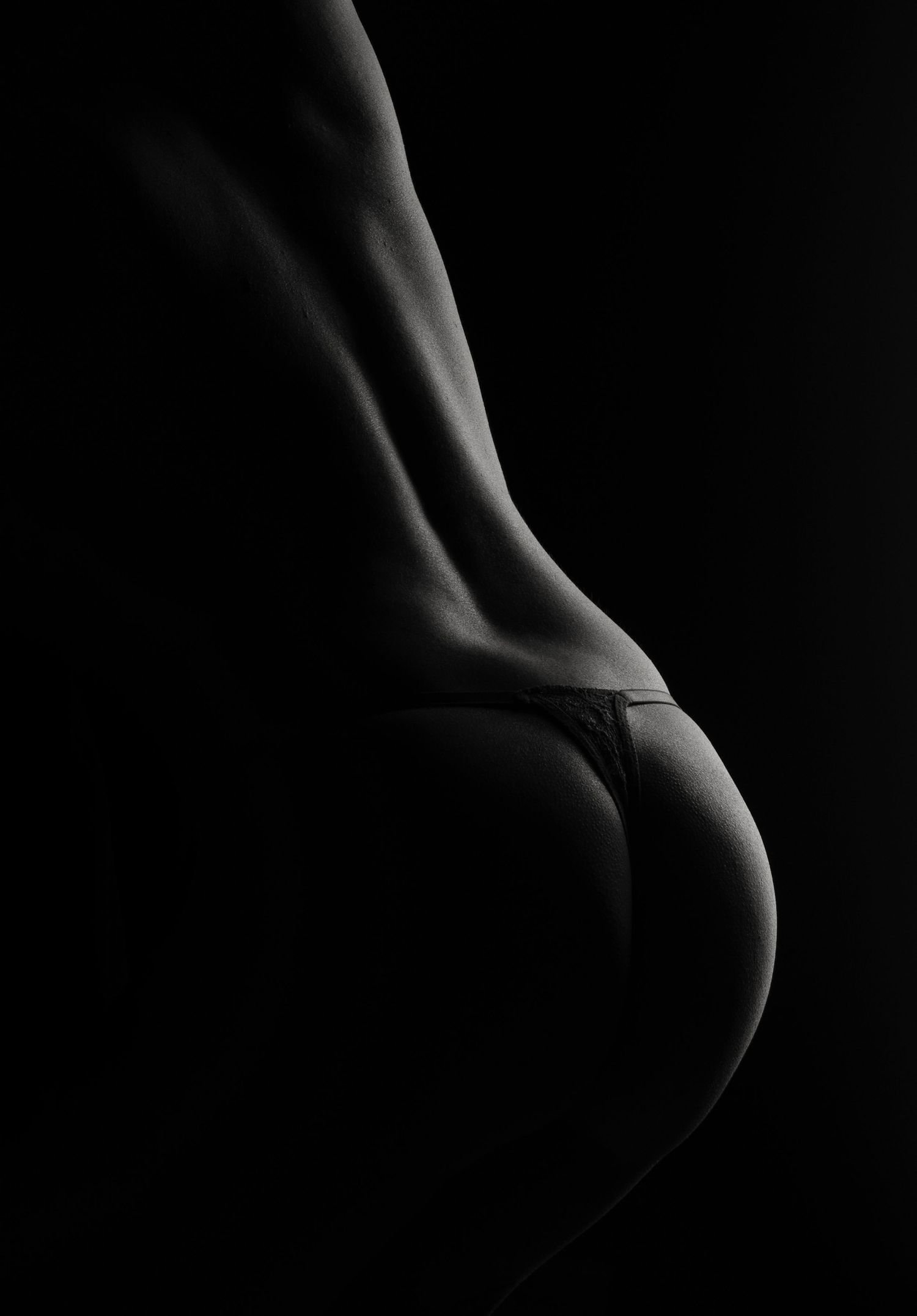 Black and white artistic photo highlighting feminine curves with dramatic low-key lighting and minimal lingerie.