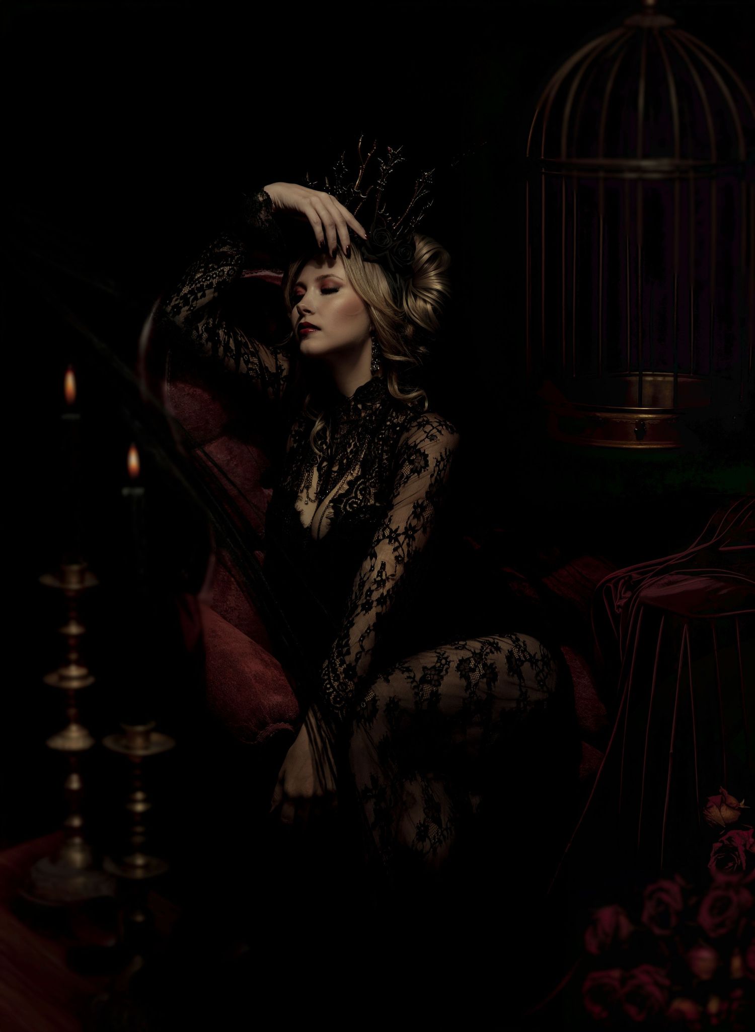 Woman in black lace gown and ornate headpiece poses with closed eyes beside lit candles, a birdcage and red roses in a dark gothic scene.