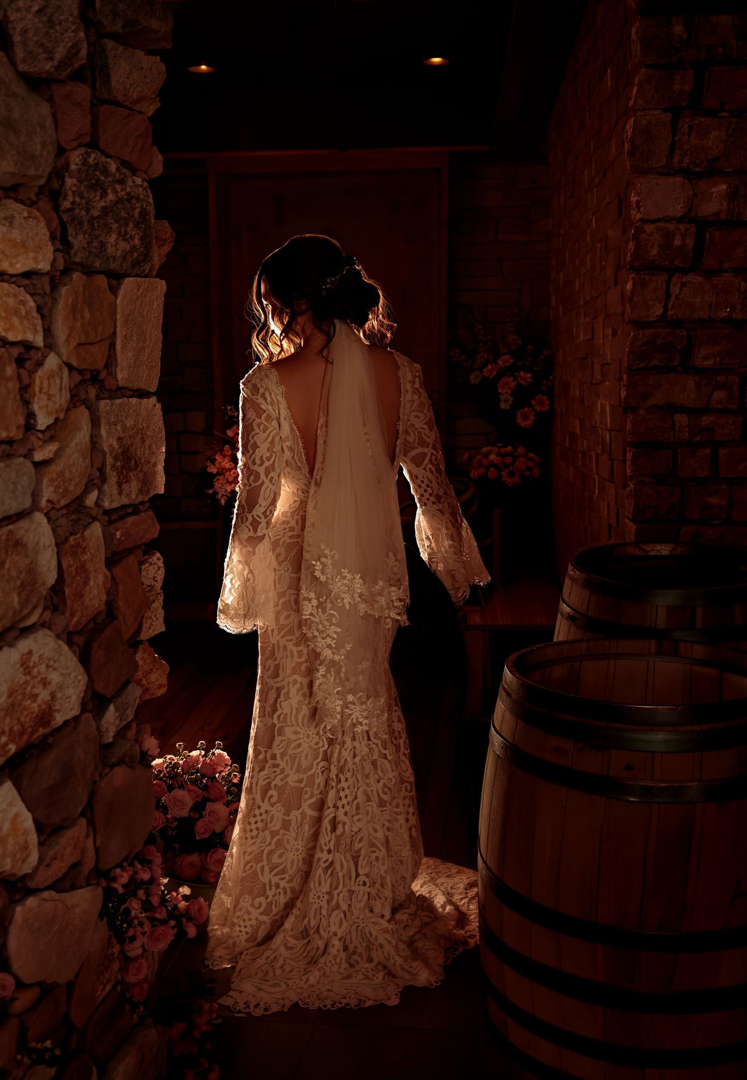 A vintage-style wedding dress with lace details is illuminated against a rustic stone wall and wooden barrels in warm lighting.