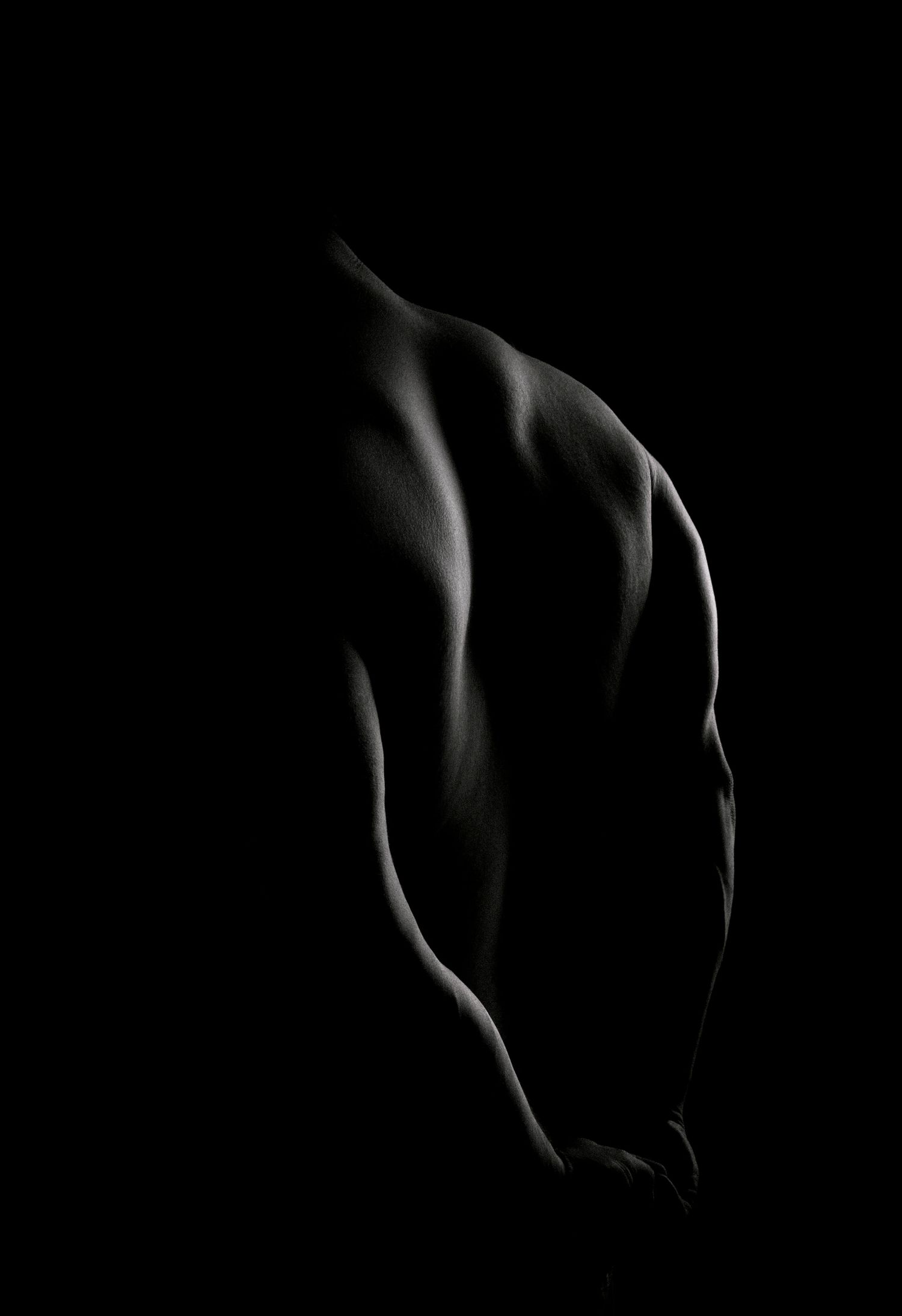 Dramatic black and white photo of a muscular male back, highlighted by low-key studio lighting against a dark background.