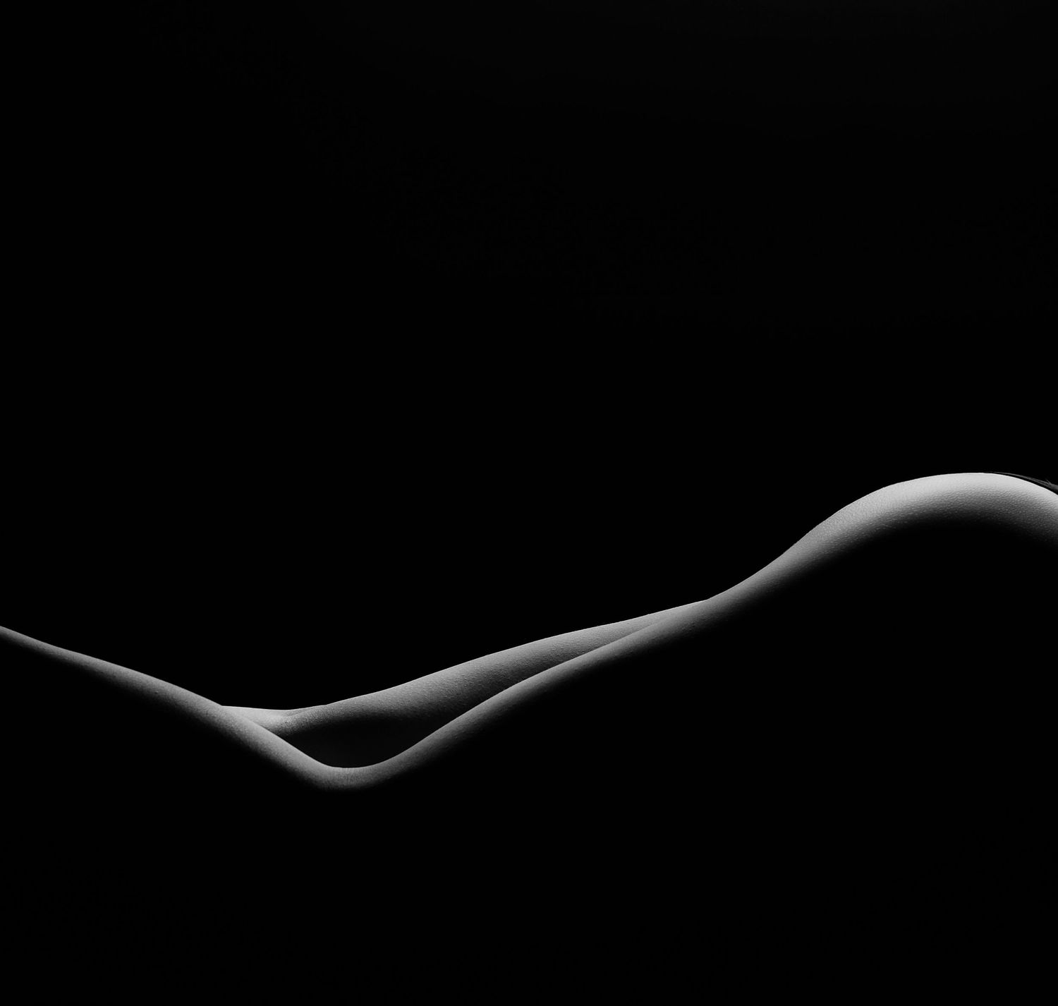 Black and white fine art nude photography showcasing elegant body curves against a dark background.
