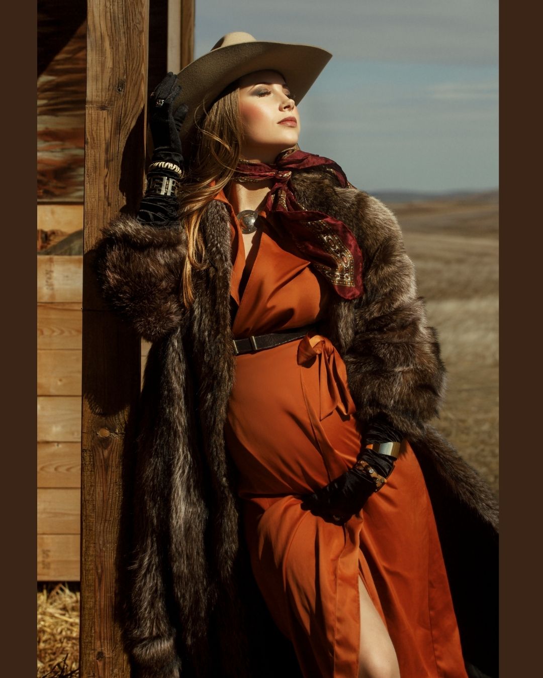 A person wearing a red hooded cape and fur collar poses dramatically against a rustic wooden backdrop at sunset.