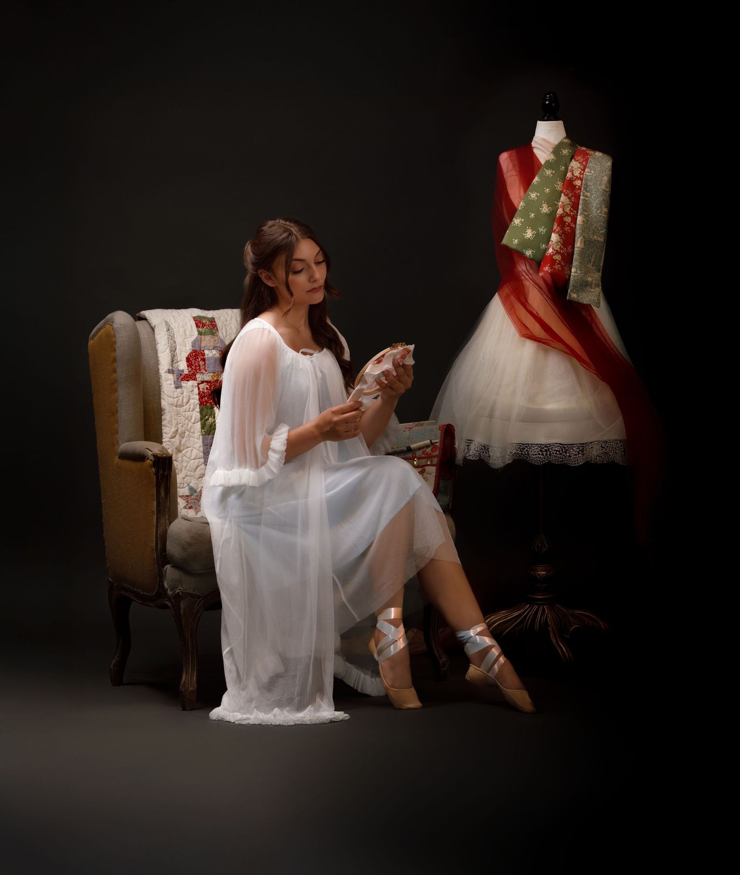 A woman in a white sheer gown sits in a vintage chair beside a mannequin displaying a colorful red and green dress.