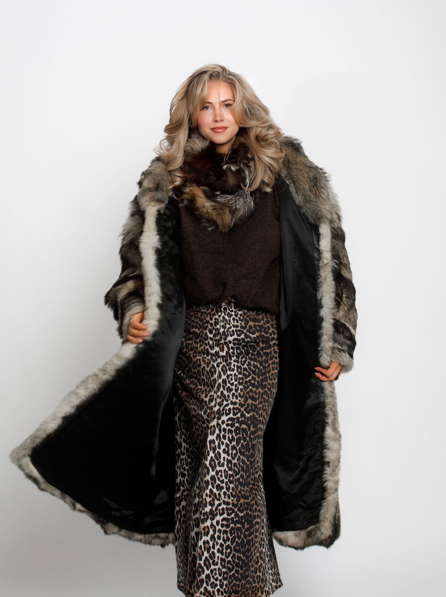 Woman modeling a long fur coat with leopard print skirt and dark top on white background.