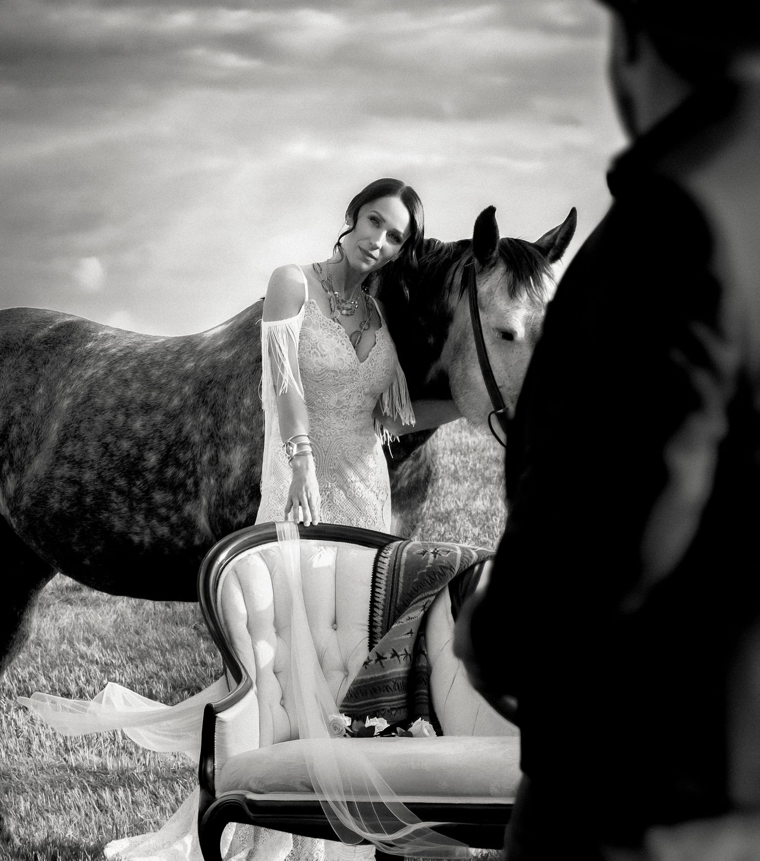 Black and white wedding photo of bride leaning on horse beside vintage chair with groom in foreground.