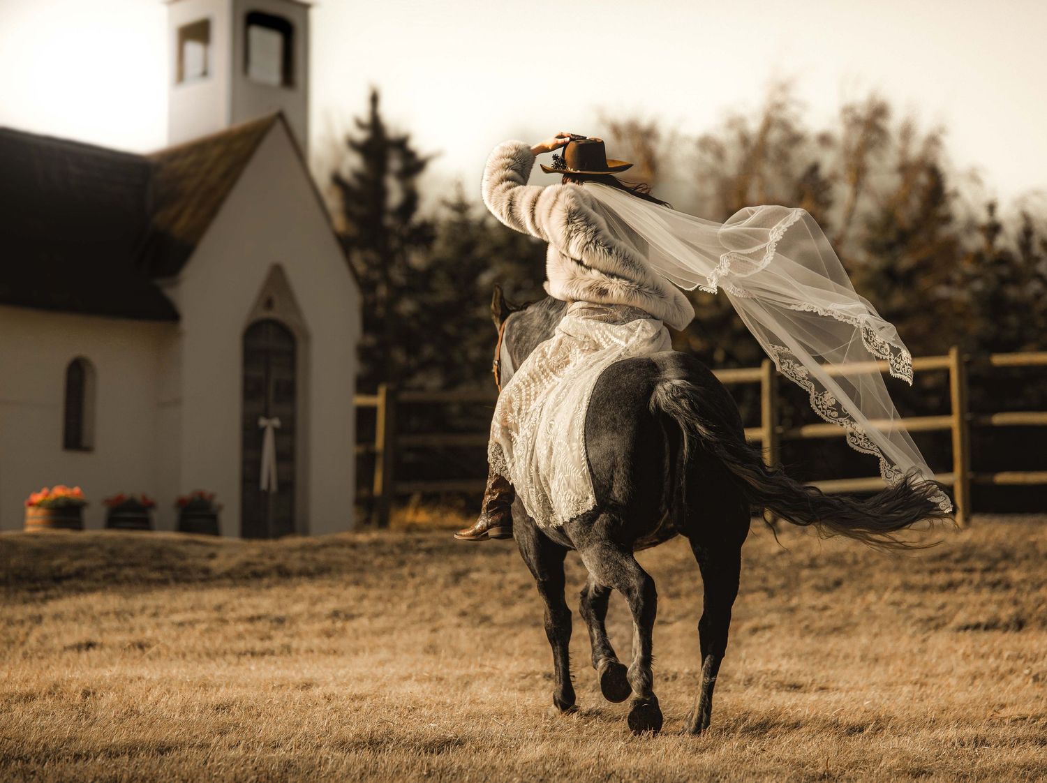 Bride in white gown and veil rides dark horse near rustic church in golden-toned outdoor wedding photo.