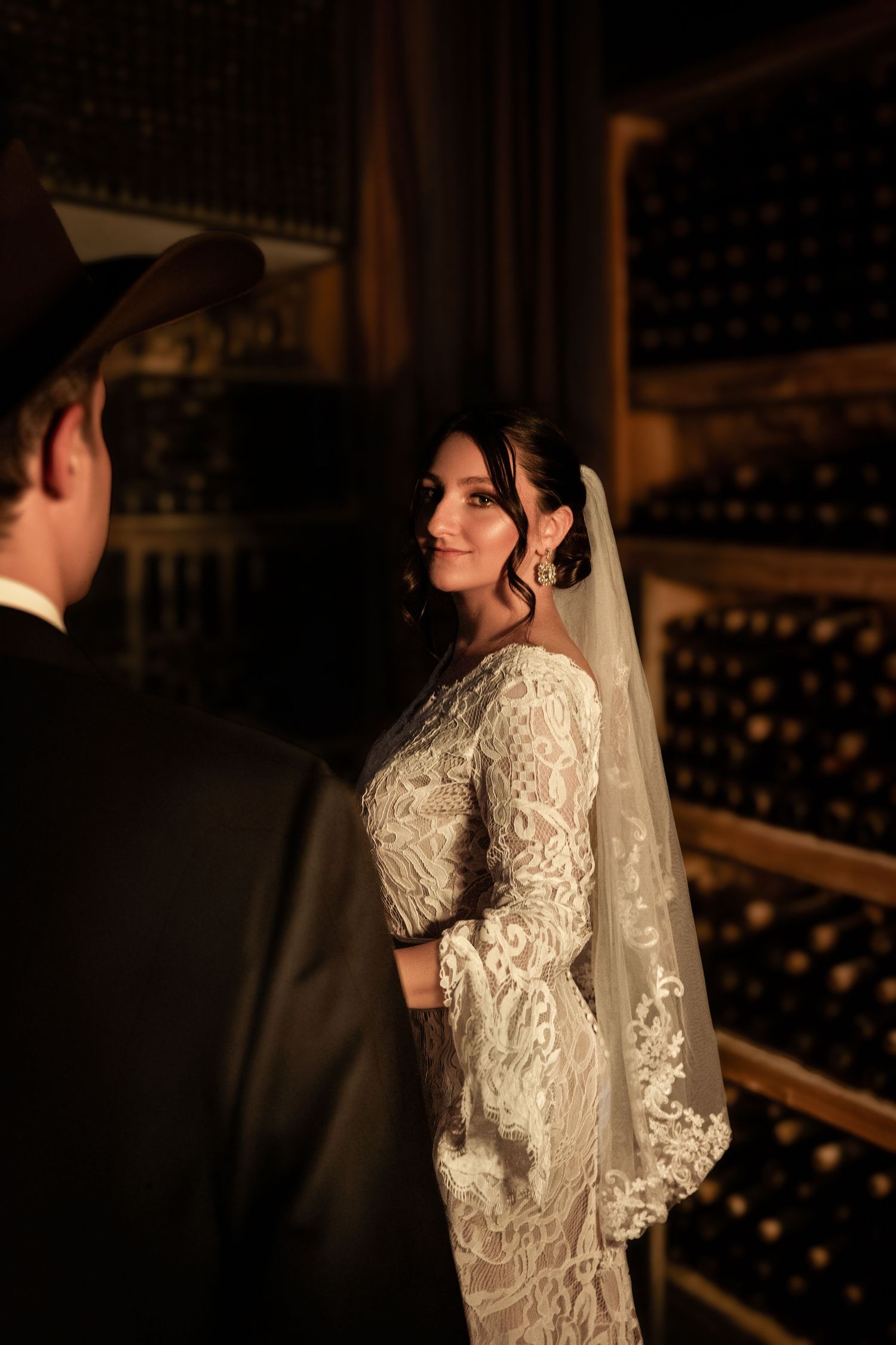 Bride in lace wedding gown and veil stands in dimly lit wine cellar, smiling at groom in dark suit.