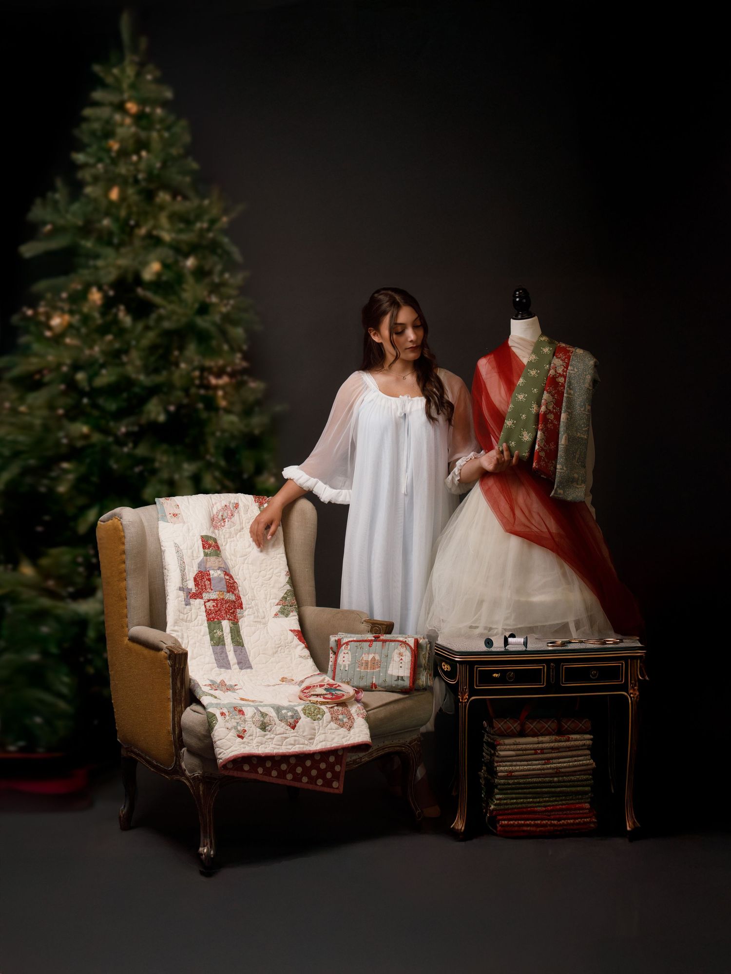 Woman in white dress poses near Christmas tree with festive quilts and decorated mannequin in holiday studio setting.