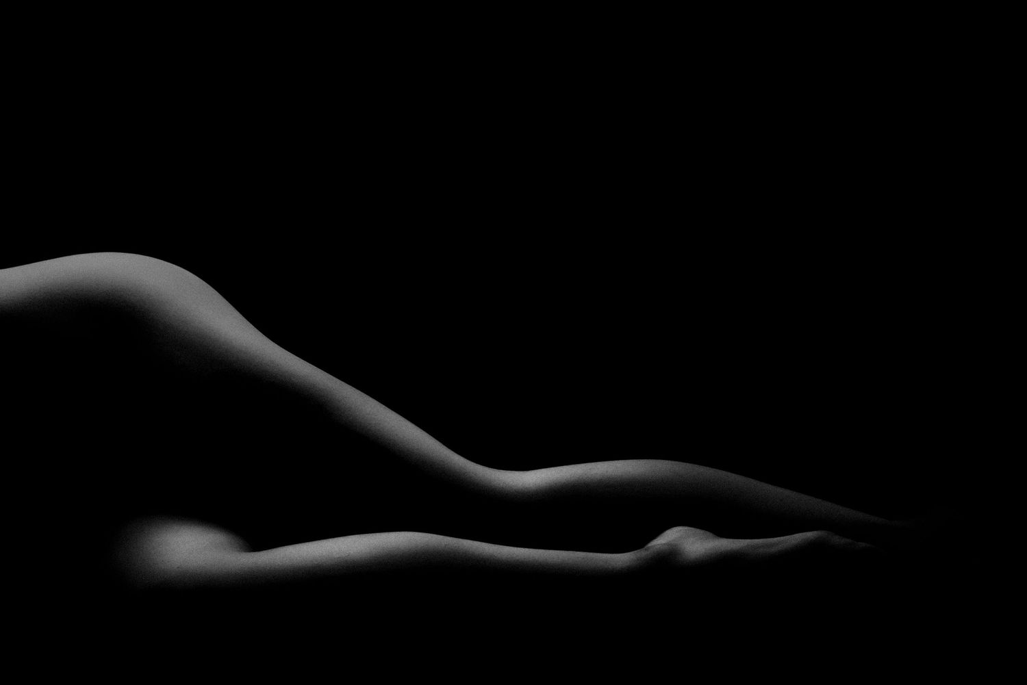 Black and white artistic nude silhouette of a reclining figure with dramatic low-key lighting on dark background.