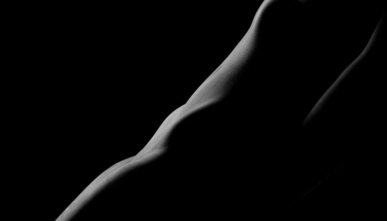Black and white artistic nude photography highlighting the curves of a human body against a dark background.