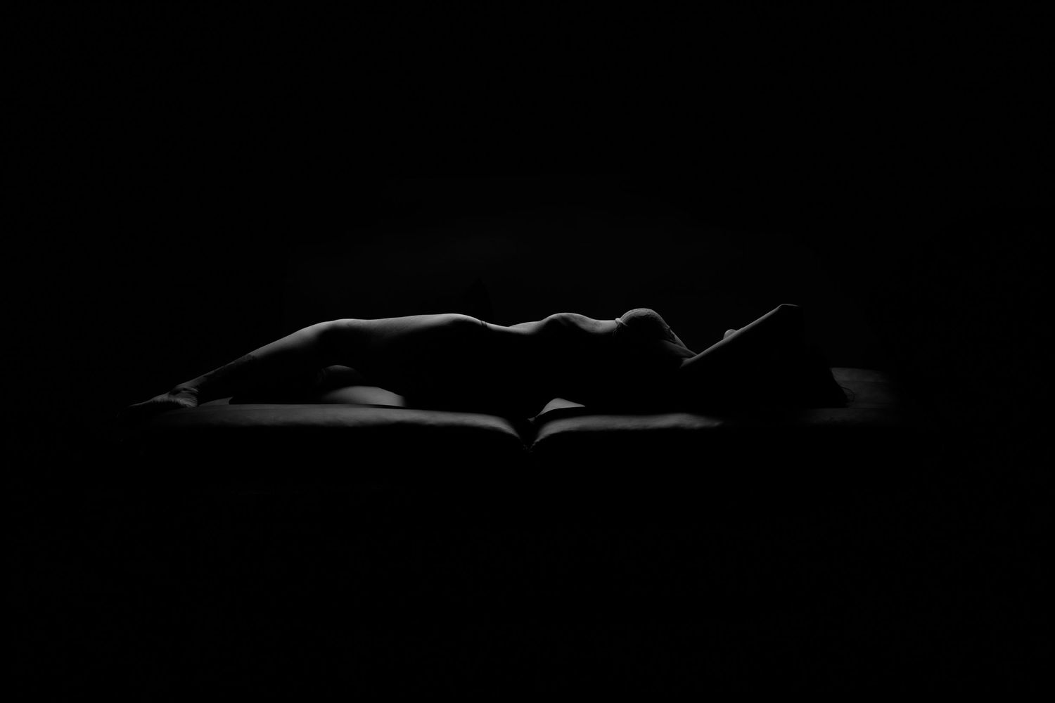 A dramatic low-key fine art nude silhouette of a figure lying down, illuminated by subtle side lighting against a dark background.