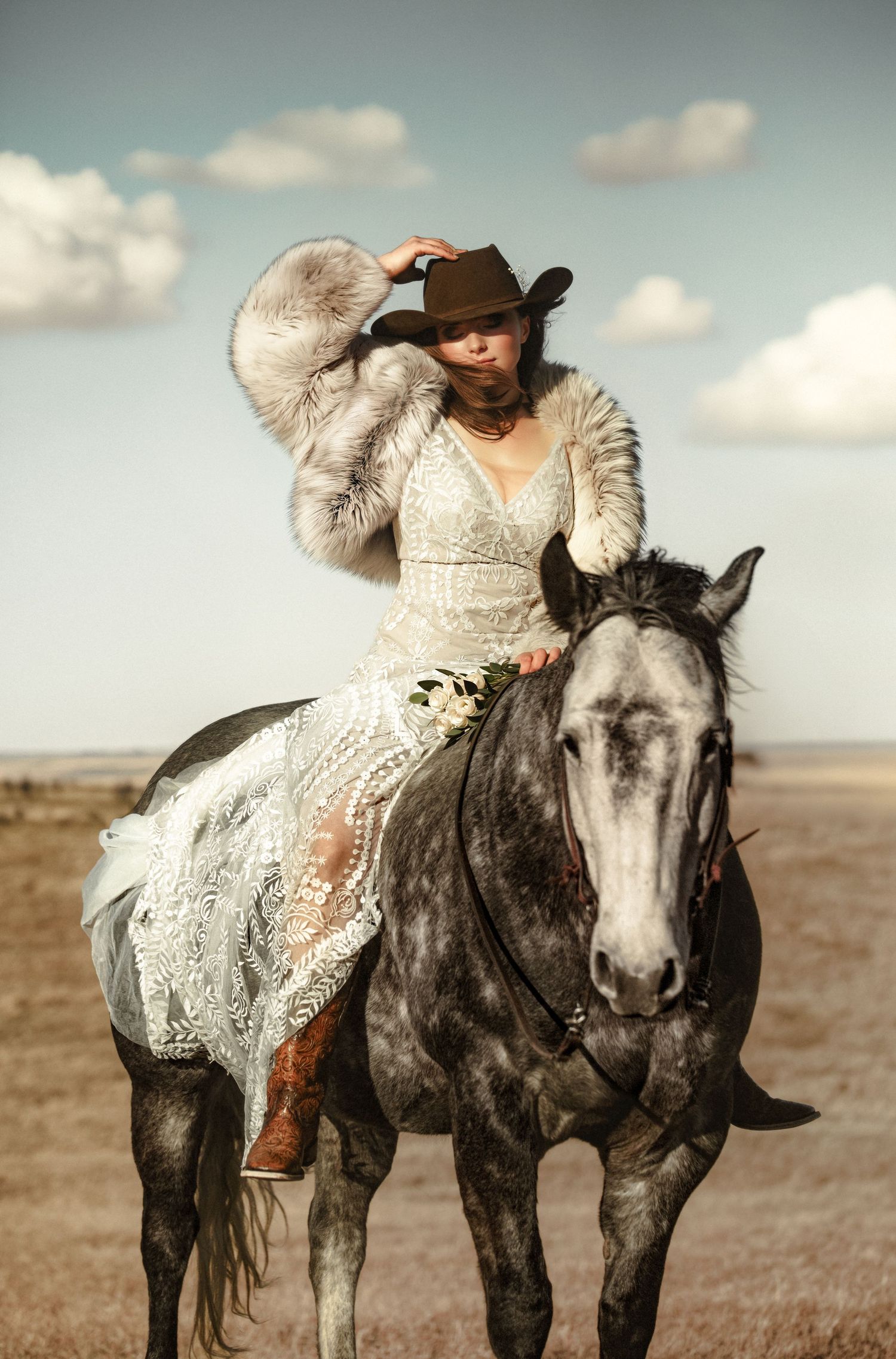 A rider in a white lace dress and brown cowboy hat sits atop a dappled gray horse against a cloudy sky backdrop.