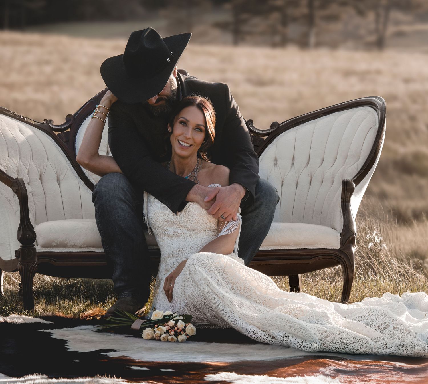 Bride in lace gown and groom in black cowboy hat share romantic moment on vintage sofa at rustic outdoor wedding.