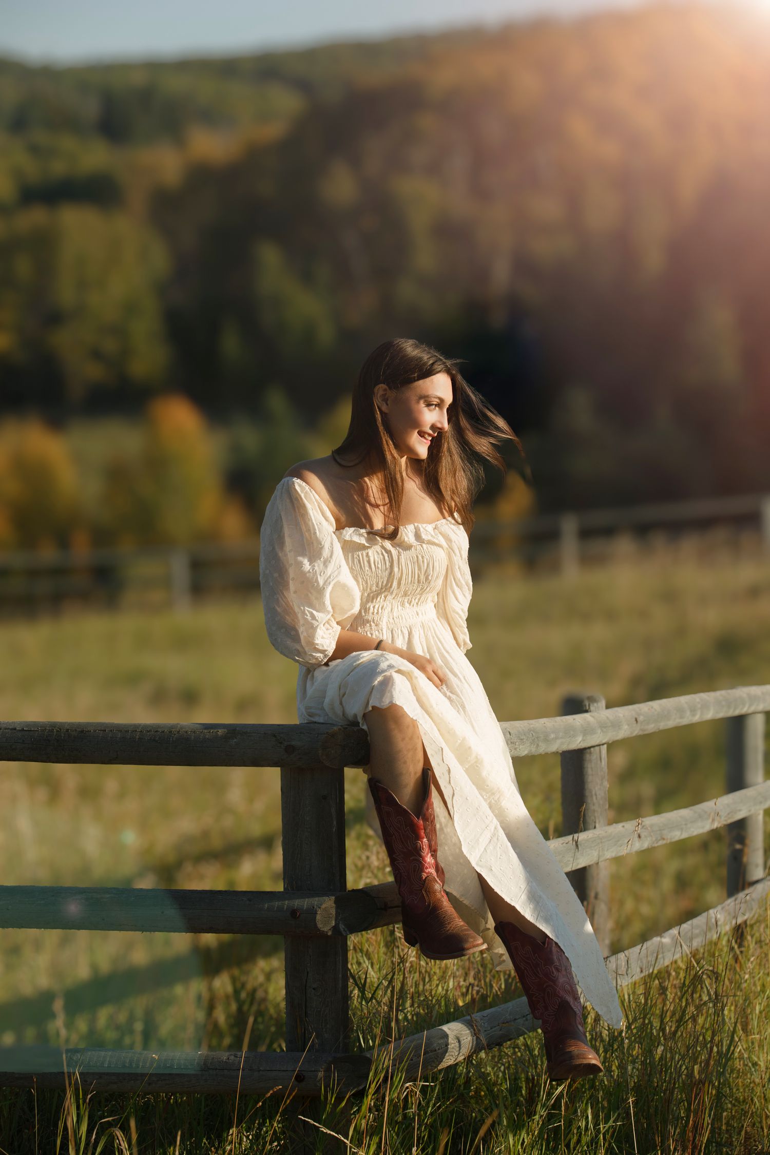 Young woman in white dress and cowboy boots sitting on wooden fence at golden hour with autumn hills backdrop.