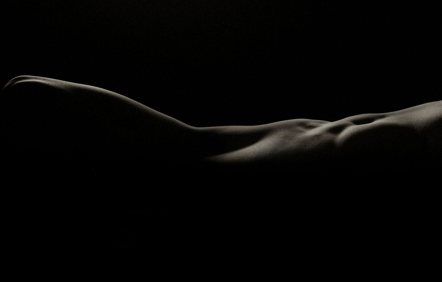 Artistic black and white nude body landscape photograph highlighting curves and contours against dark background.