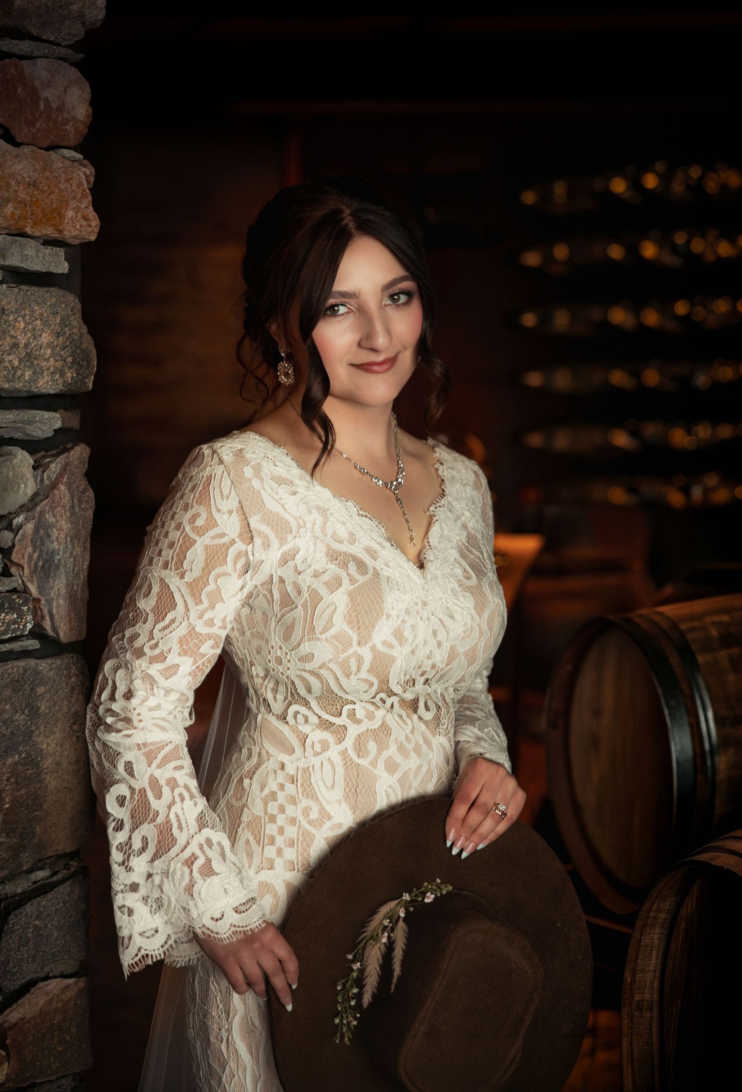 Woman in white lace dress holding a brown hat poses elegantly in a rustic wine cellar with barrels.