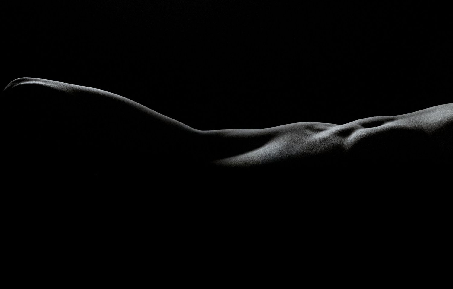 Black and white artistic nude bodyscape photography highlighting curves and contours against dark background.