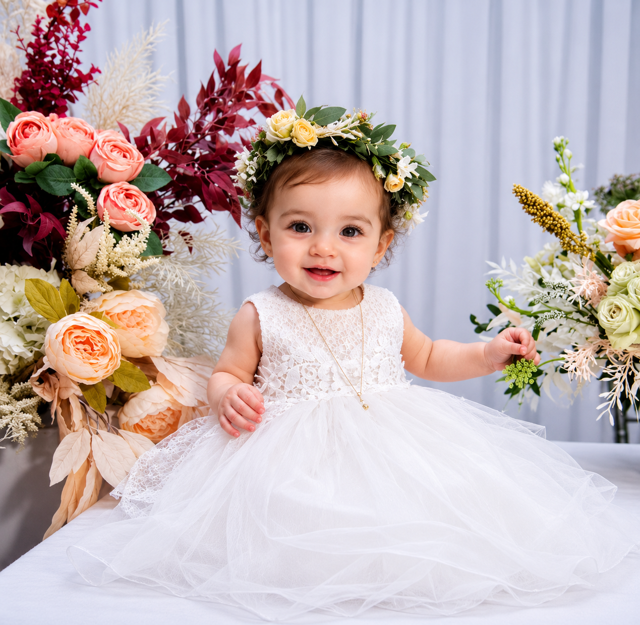 Adorable baby in white dress and floral crown poses between colorful bouquets at a baptism celebration.