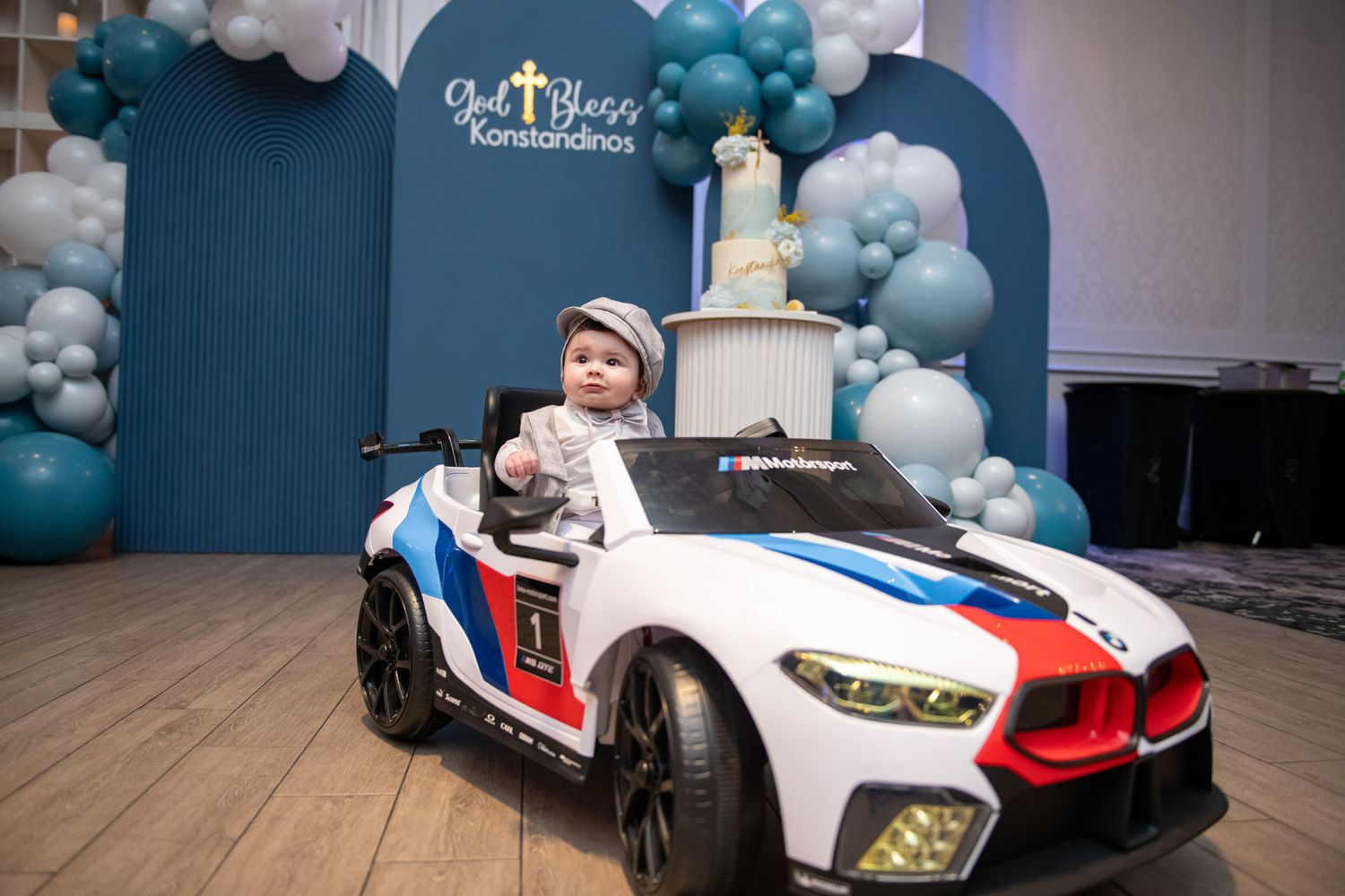 Child portrait at a Greek Orthodox baptism. Smiling baby boy in white cap seated in toy BMW car at baptism celebration with balloon decor.