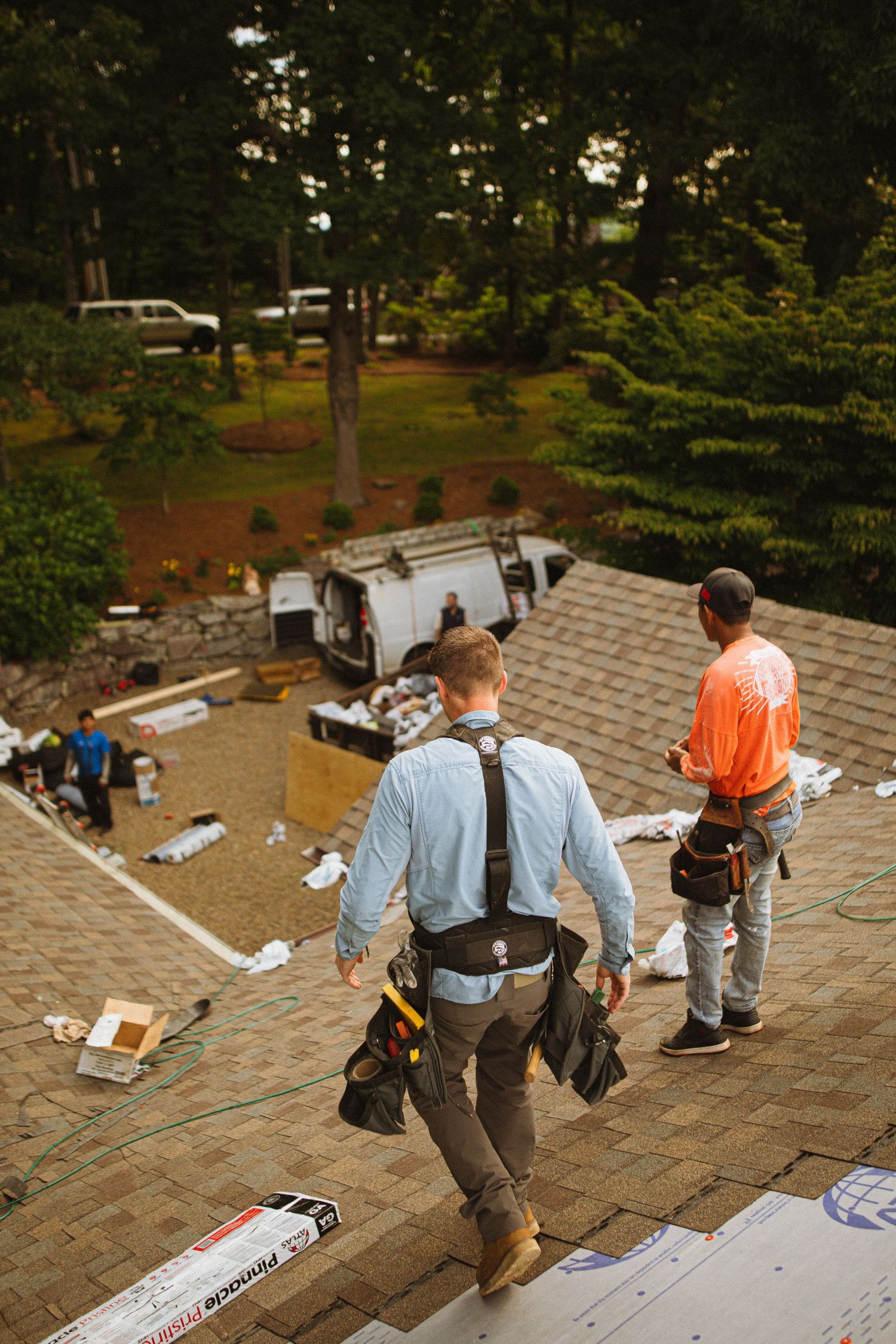 Two roofers wearing tool belts walk across a shingled roof during a roofing installation project.
