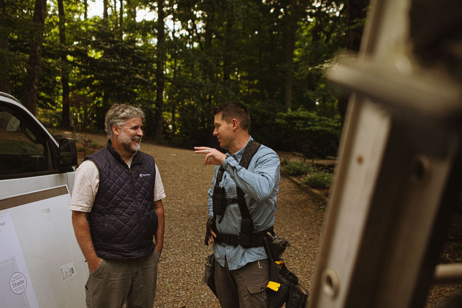 Two men having a conversation outdoors near a vehicle, surrounded by lush green trees in a wooded setting.
