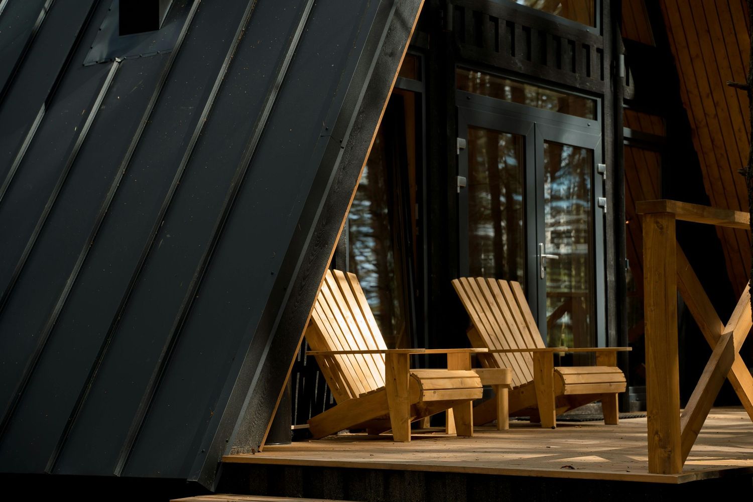 Two wooden Adirondack chairs sit on a sunlit deck of an A-frame cabin with dark metal roofing.