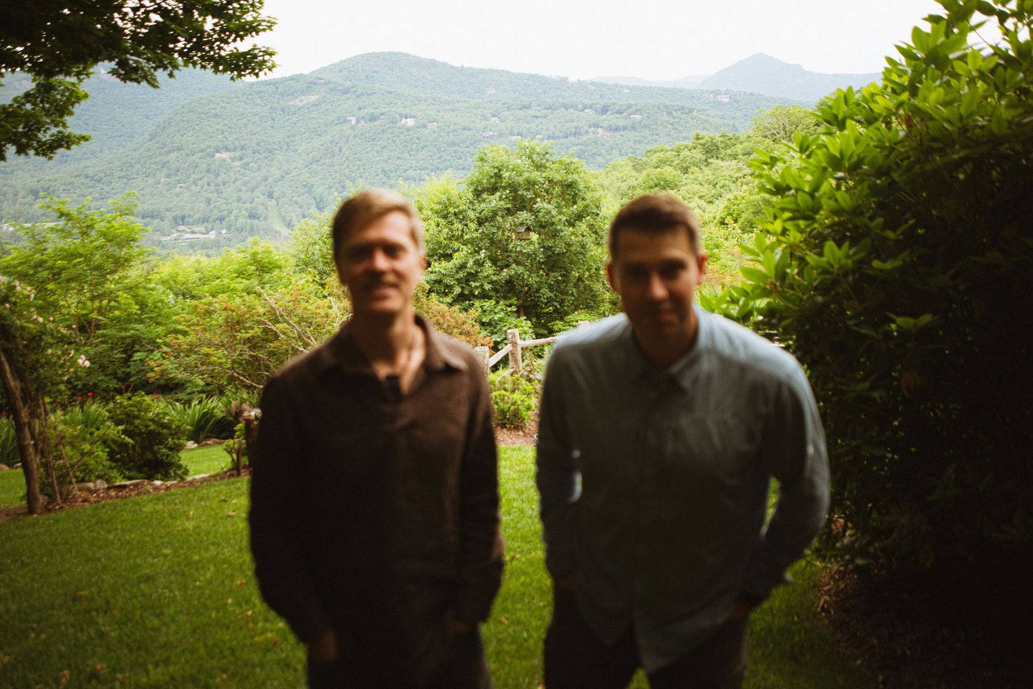 Two men standing outdoors on a green lawn with lush trees and mountain landscape in the background.
