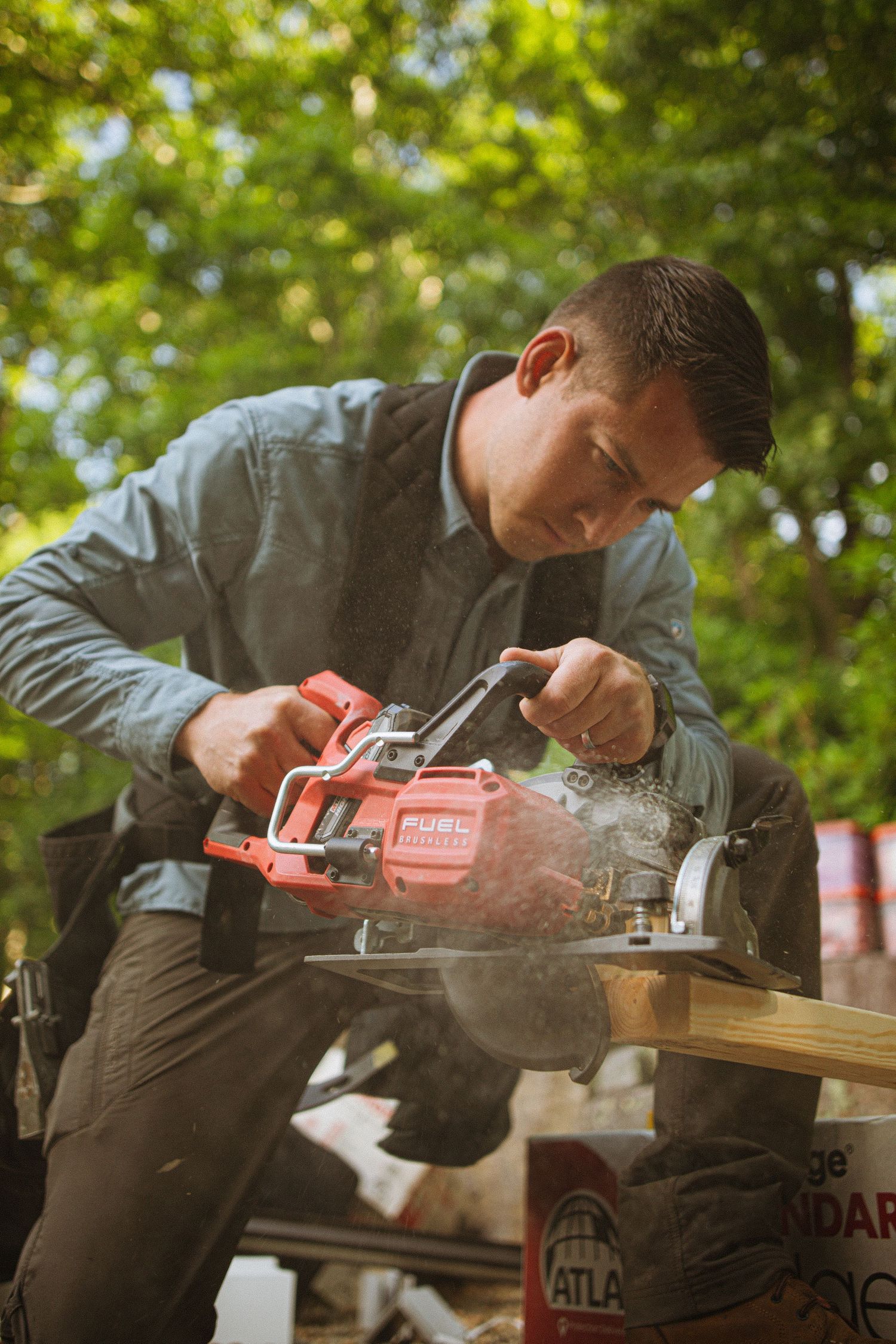 Man using a red FUEL circular saw to cut wood outdoors, with sawdust flying and green trees in background.