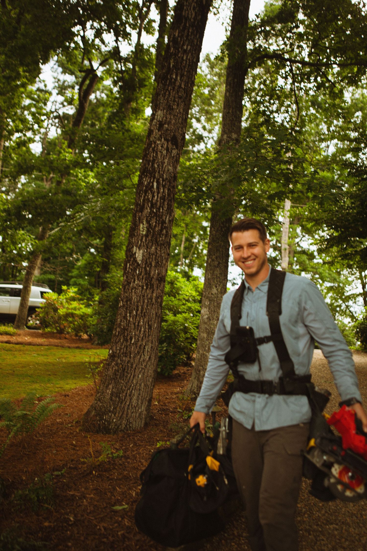 Smiling man carrying gear bags walks through a lush green wooded area near a lake.