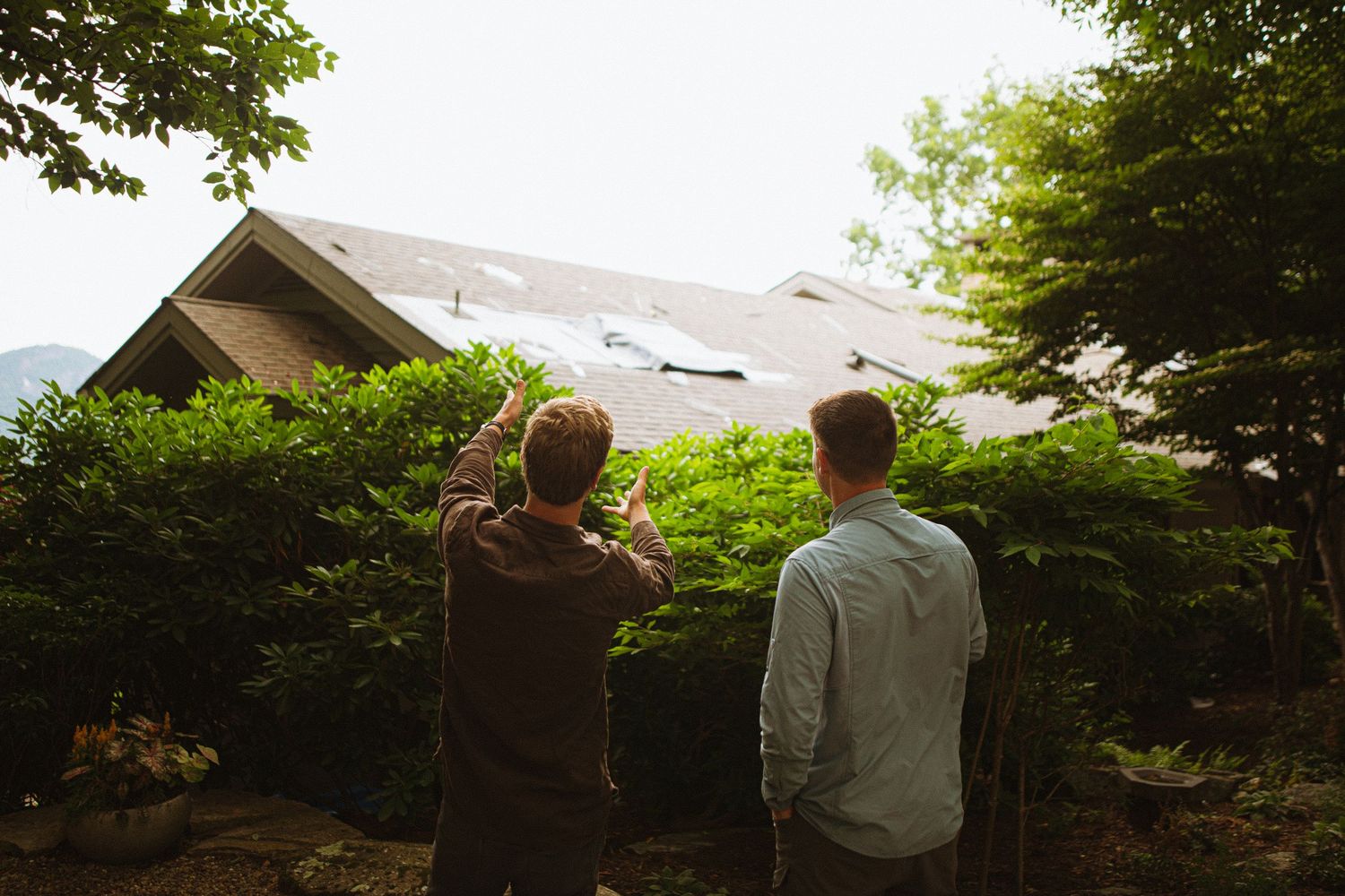 Two men stand with backs turned examining a house roof surrounded by lush green trees and shrubs outdoors.