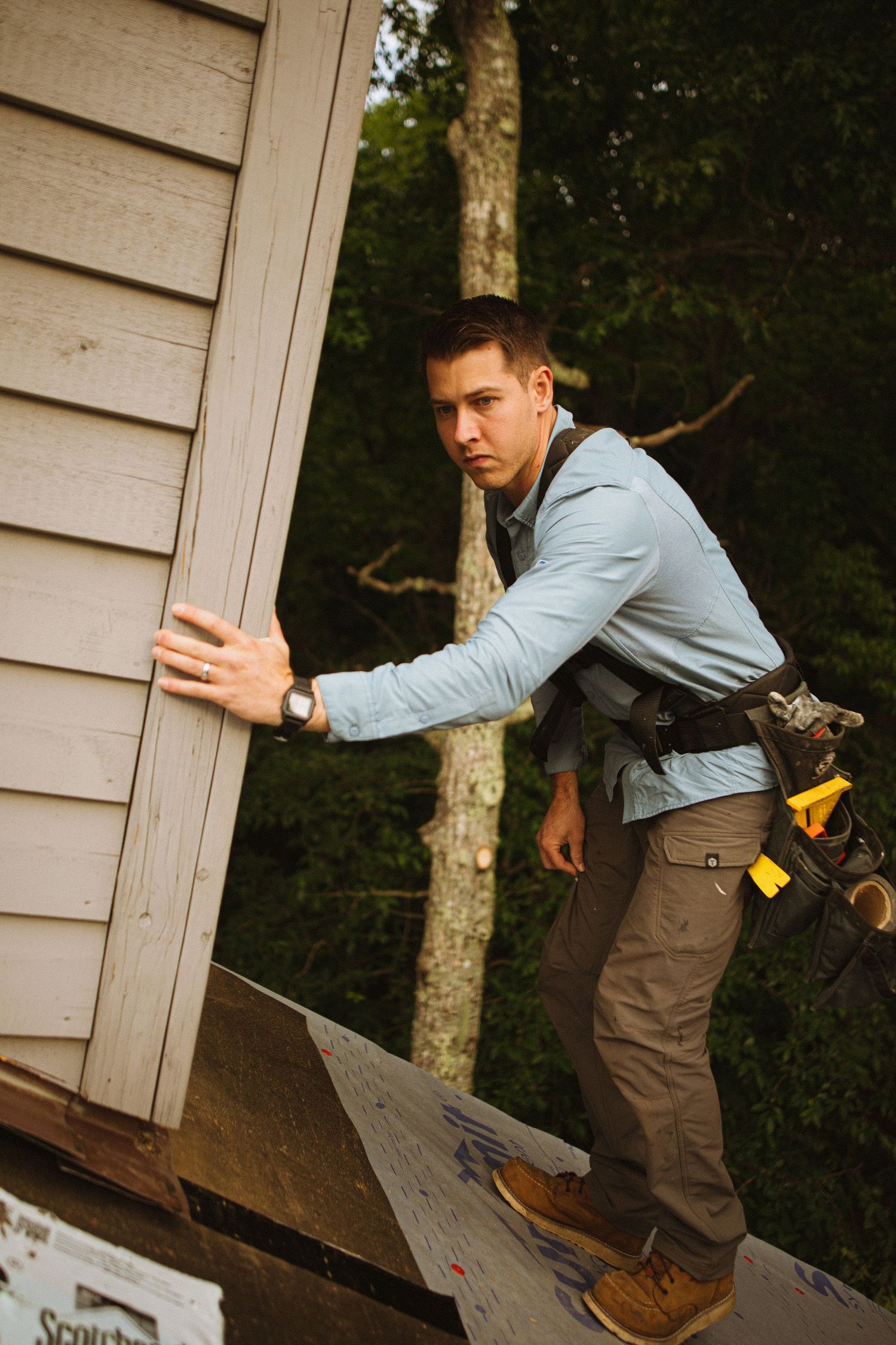 Young male contractor in tool belt installs siding on a house roof, wearing a blue shirt and work boots outdoors.