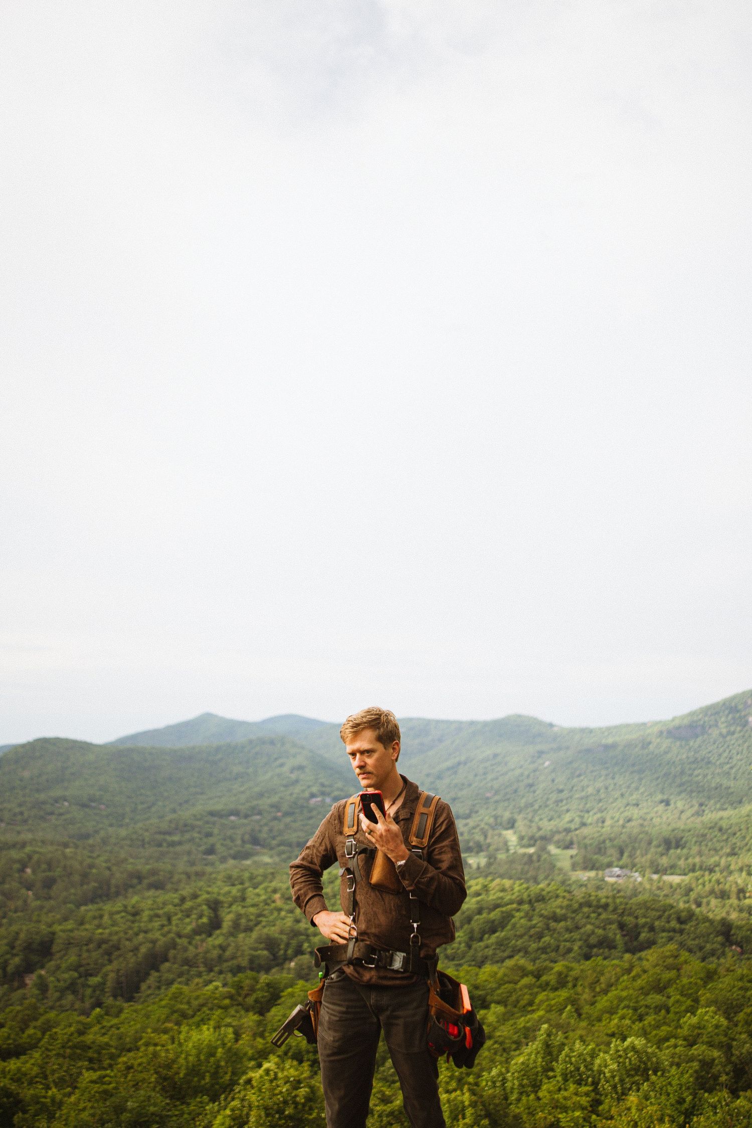 Man with camera gear checks phone on mountain overlook with lush green forested hills in background.