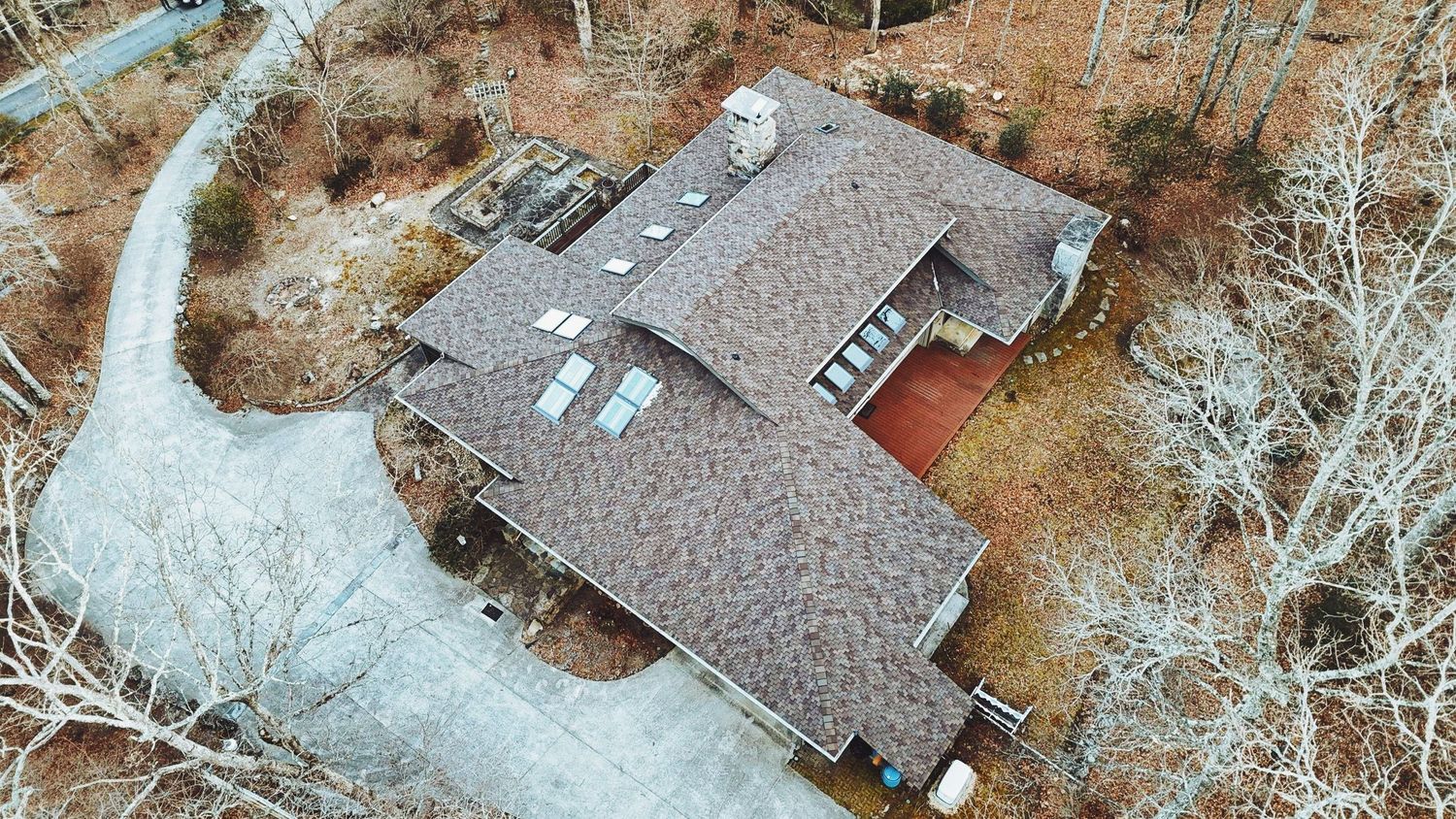 Aerial drone view of a large residential home with brown shingle roof surrounded by bare trees in a wooded setting.