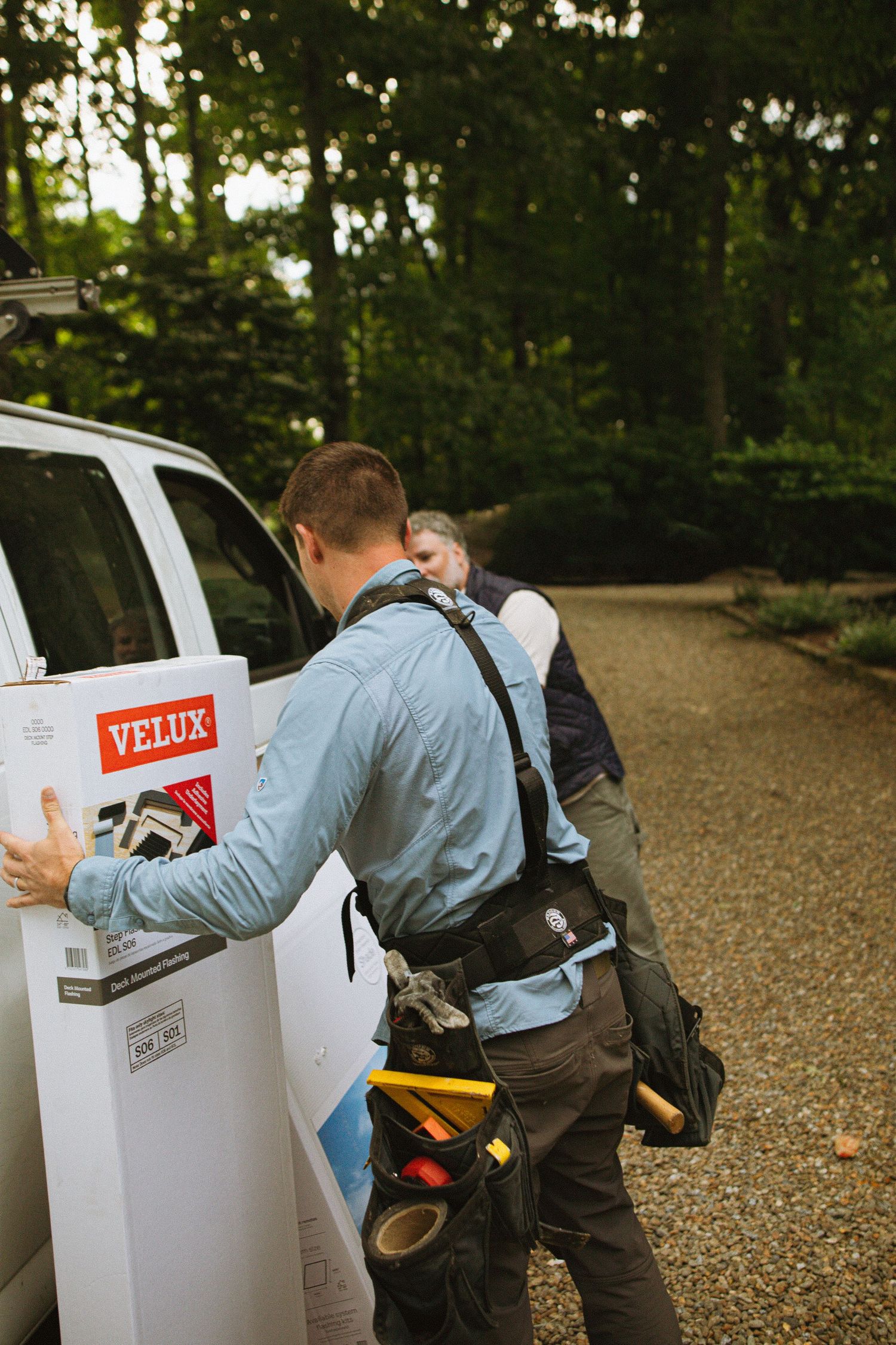 Two workers unloading a Velux skylight box from a white van in a tree-lined driveway.
