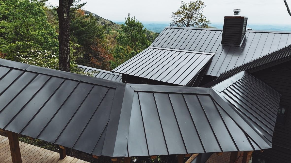 Dark gray standing seam metal roof on a cabin surrounded by lush trees with scenic mountain views in the background.