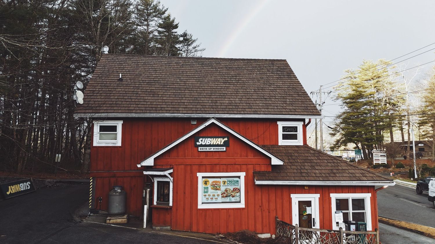Red barn-style building with white trim serves as a small shop, surrounded by trees with a rainbow visible in the cloudy sky.