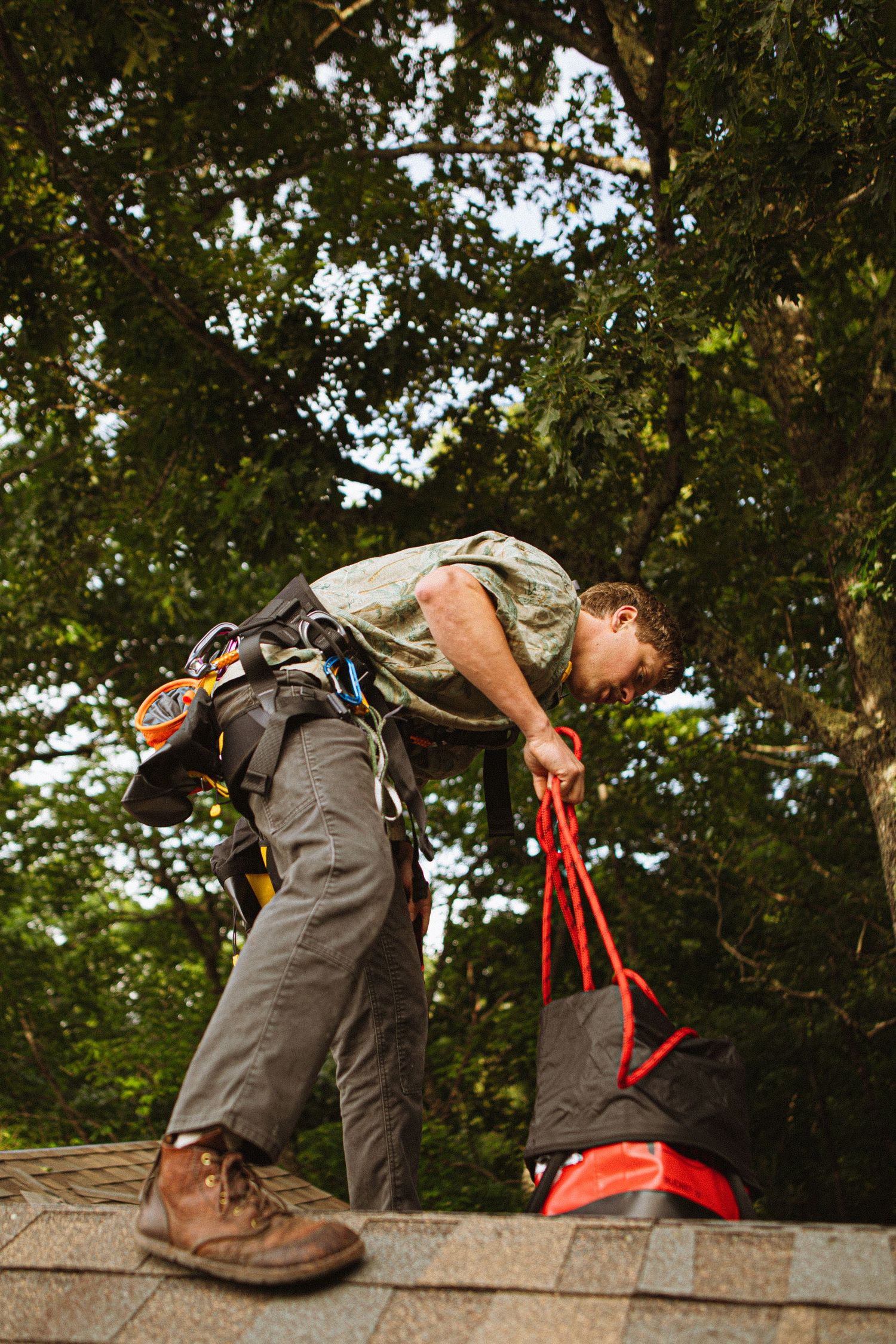 A roofer in safety harness and work boots bends over equipment on a shingled roof with trees in background.