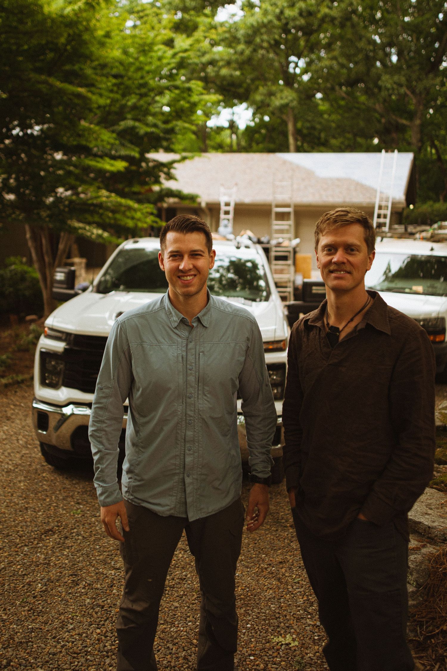 Two men smiling outdoors near trucks and a house with ladders, surrounded by lush green trees.
