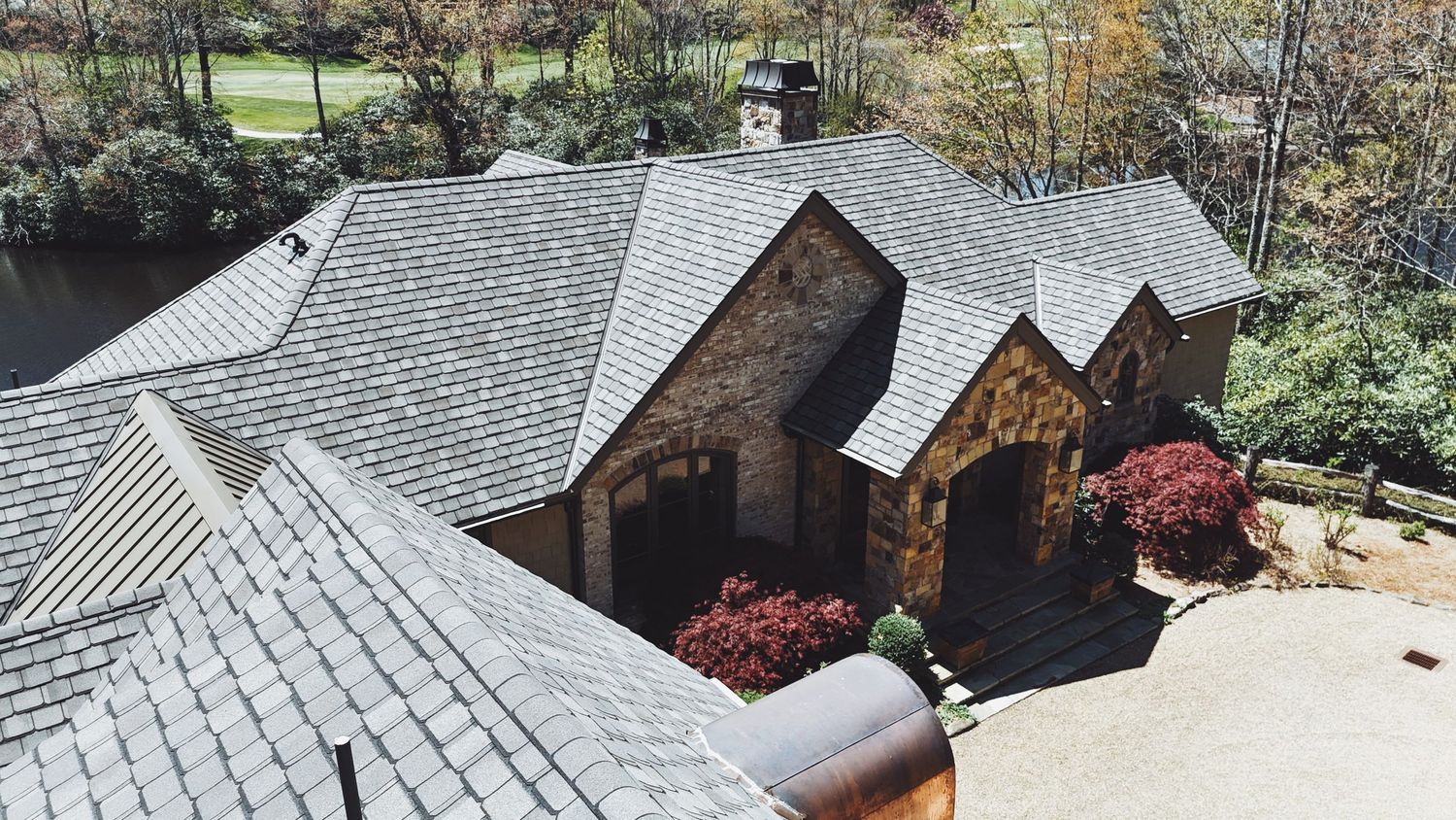 Aerial view of a stone cottage home with gray slate roof, surrounded by lush trees and colorful landscaping.