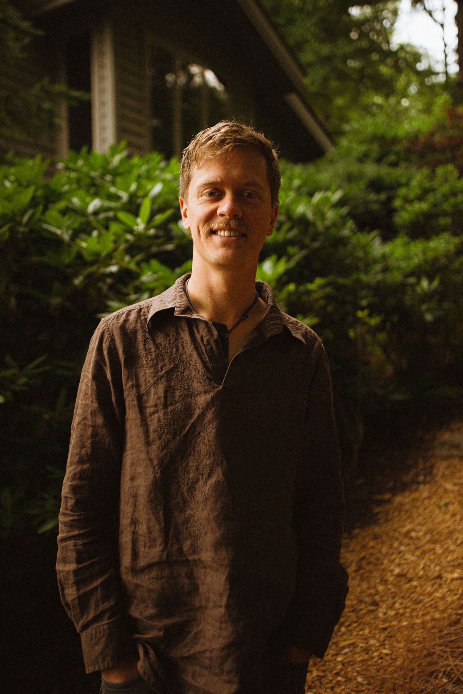 A smiling man in a brown linen shirt stands on a garden path surrounded by lush green foliage near a wooden building.