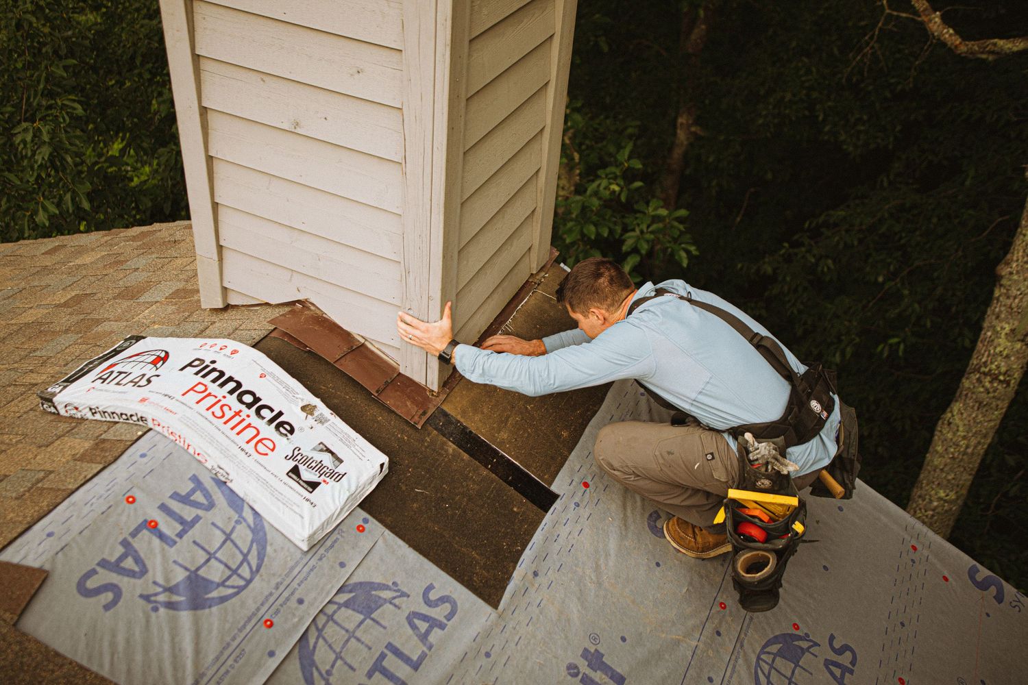 Roofer installing shingles near chimney flashing on residential roof with Atlas Pinnacle roofing materials nearby.