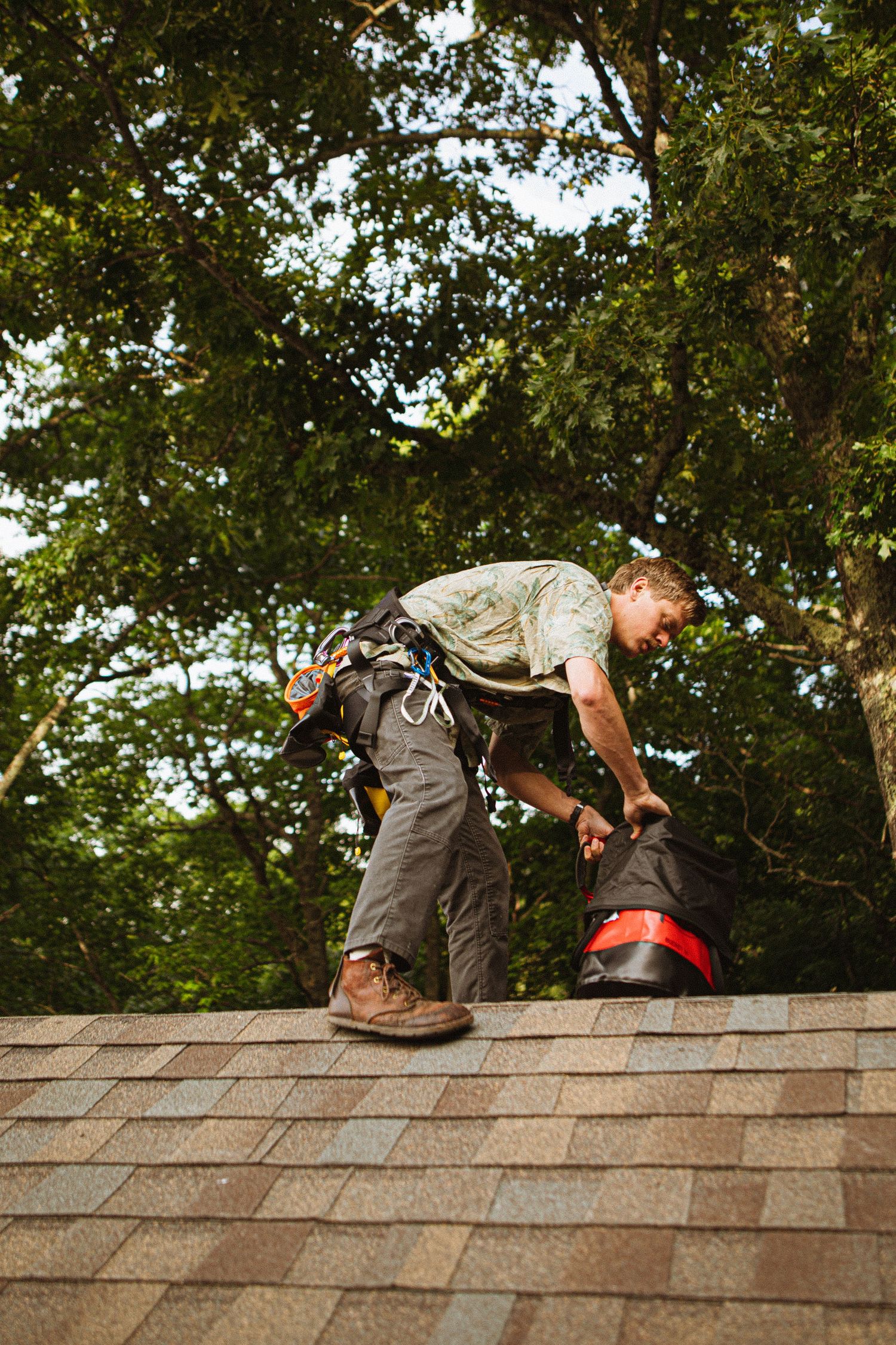 Roofer in safety harness working on shingled roof surrounded by green trees.