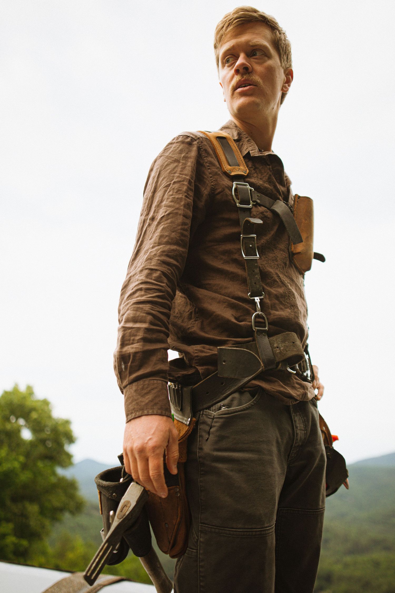 A rugged man in tactical harness holds a gun outdoors with mountain greenery behind him.